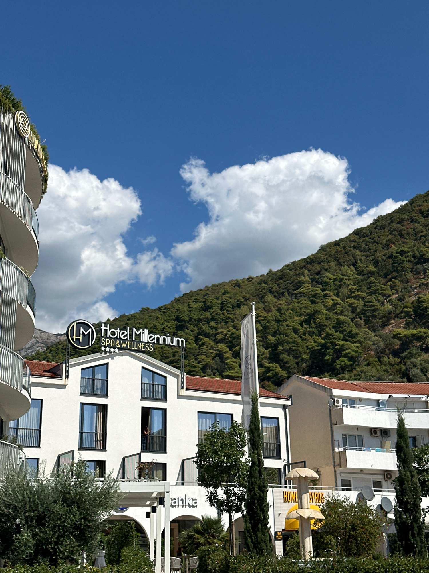 The image shows the Hotel Millennium in Budva, Montenegro. The hotel is a white building with a red-tiled roof. There are balconies on each floor, and a sign that says "Hotel Millennium Spa & Wellness." In the background, there is a green mountain covered in trees. The sky is blue with some white clouds. In front of the hotel, there are some trees and bushes. To the left of the hotel, there is another building with a curved facade and balconies. To the right of the hotel, there is a building with white balconies and air conditioning units. There is also a street sign that says "HOME BYROS."