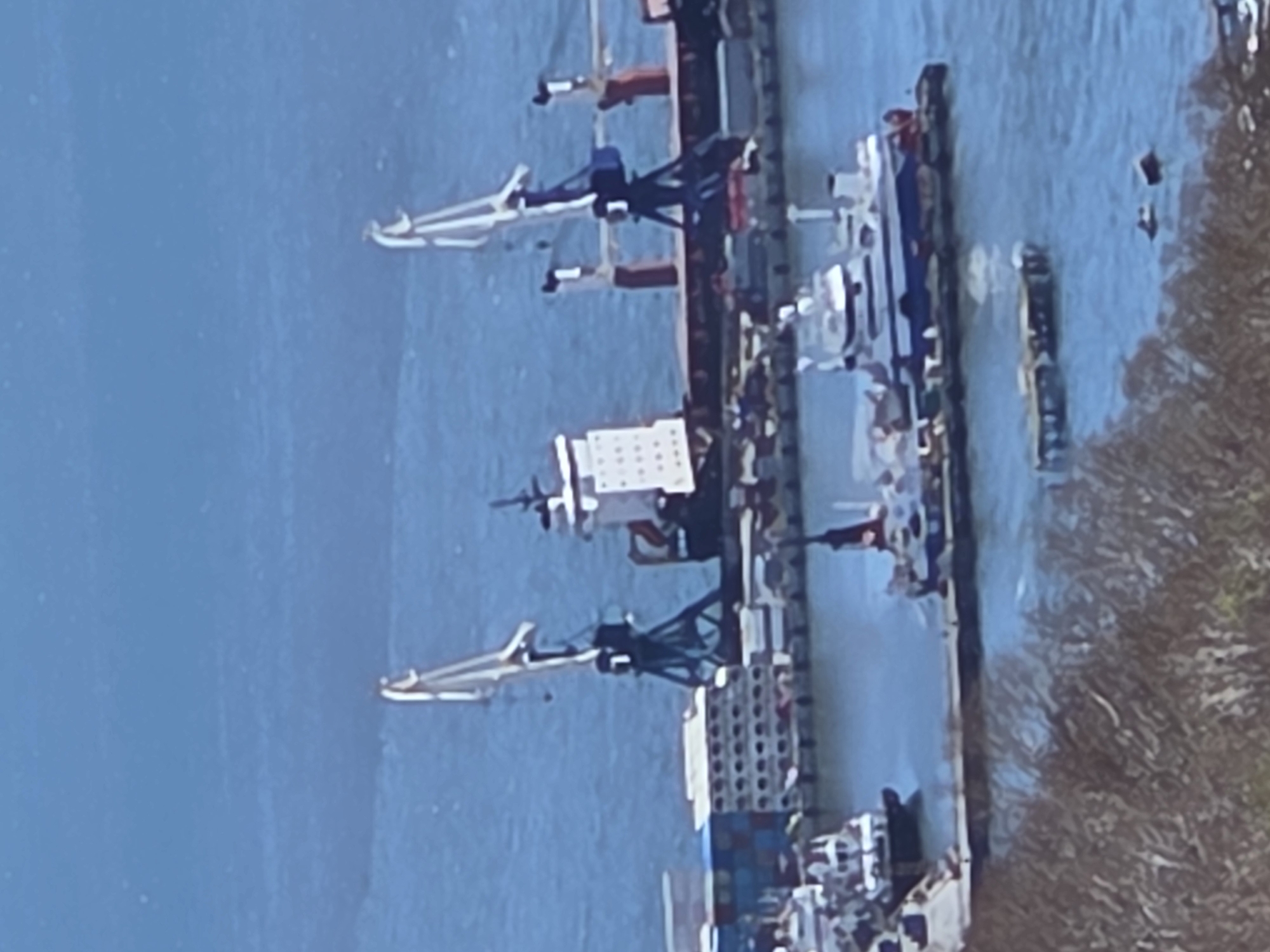 The image depicts a cargo ship docked at a port. The ship is equipped with cranes for loading and unloading containers. The port is located in a body of water. The environment appears to be partly natural. The location is in the city of Unknown, Unknown.