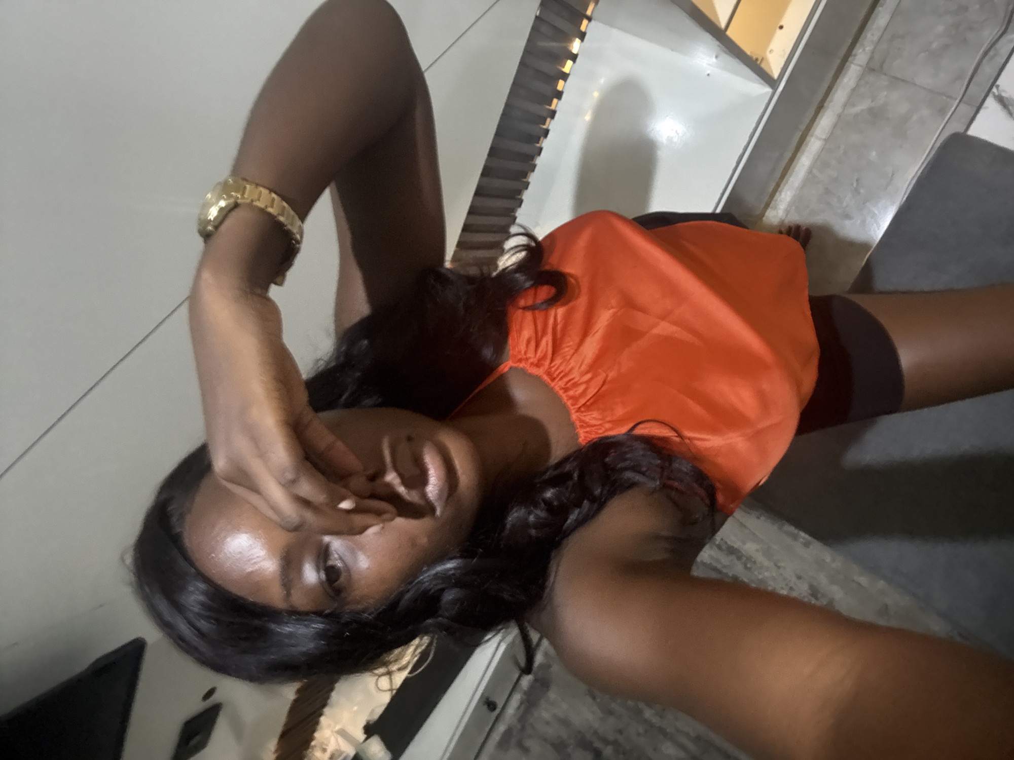 A woman is lying on her back. She is wearing an orange top, black shorts, and a gold watch on her left wrist. Her right hand is covering part of her face. The location appears to be indoors, possibly on a floor near stairs. The city is Iddo Tudunwada, Nigeria.