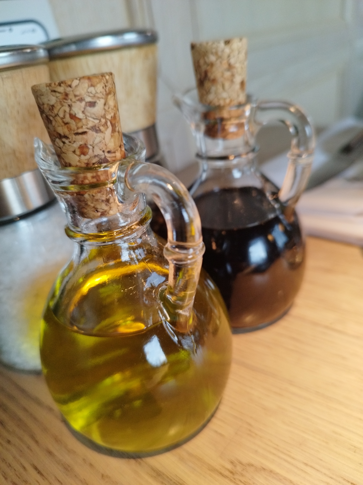 Two glass cruets with cork stoppers are placed on a wooden surface. The cruet in the foreground contains a golden-yellow liquid, likely olive oil, which reflects light. The cruet in the background contains a dark liquid, possibly balsamic vinegar. Behind the cruets, out of focus, are two cylindrical containers with metal and wooden accents, possibly spice grinders or dispensers. The lighting suggests an indoor setting, possibly a kitchen or dining area. There are no people or animals present in the image. The image is taken from a slightly elevated angle, focusing on the cruets. The scene is set on a wooden countertop. The location of this image in Saint Petersburg, Russia, cannot be determined from the visual information provided.