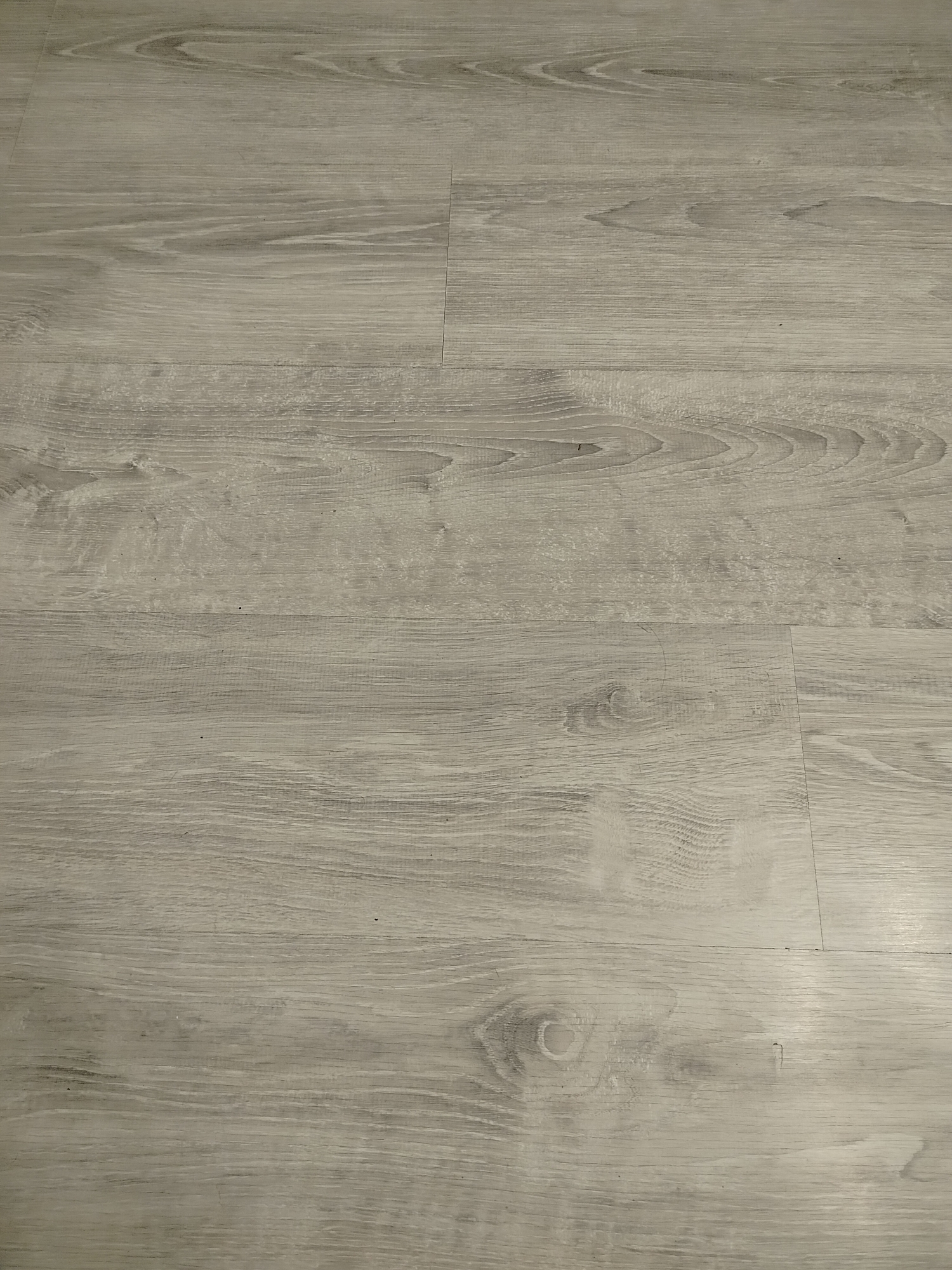 The image shows a close-up of light gray wood-look flooring with visible seams between planks. The flooring exhibits a wood grain pattern. The location is not identifiable from the image. There are no people, subjects, objects, actions, or interactions visible other than the flooring itself.