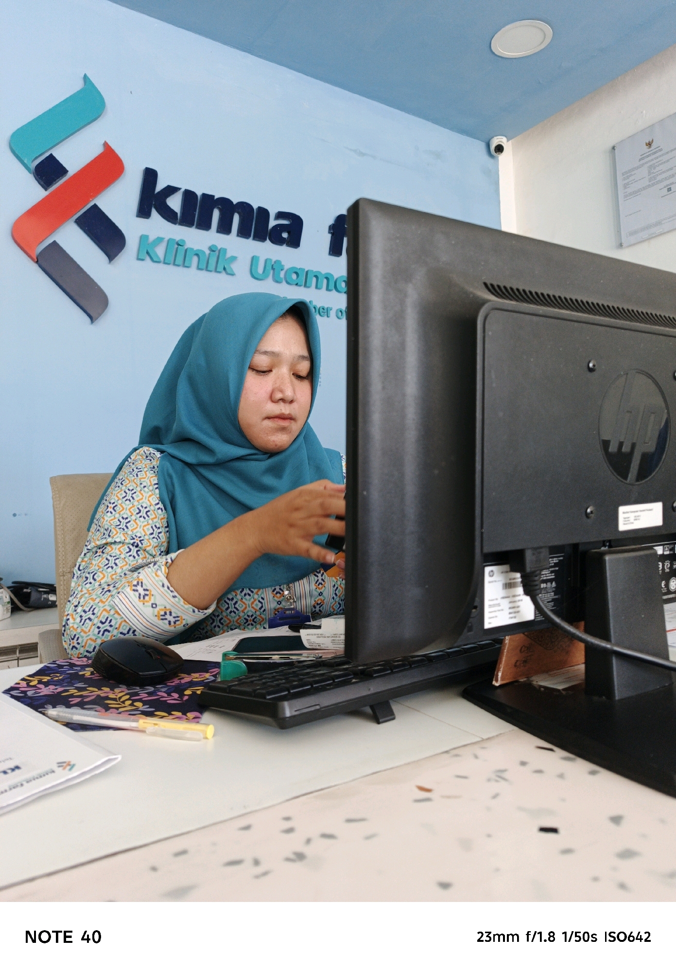 A woman wearing a teal hijab and a patterned shirt sits at a desk in front of a computer monitor. The woman is looking down at something on the desk, and her right hand is extended towards it. The computer monitor is black and has an HP logo on the back. On the wall behind the woman, there is a sign with the words "Kimia" and "Klinik Utama" written on it. A small security camera is visible on the ceiling. The desk has a keyboard, mouse, and some papers on it. The overall environment appears to be an office or a clinic reception area.