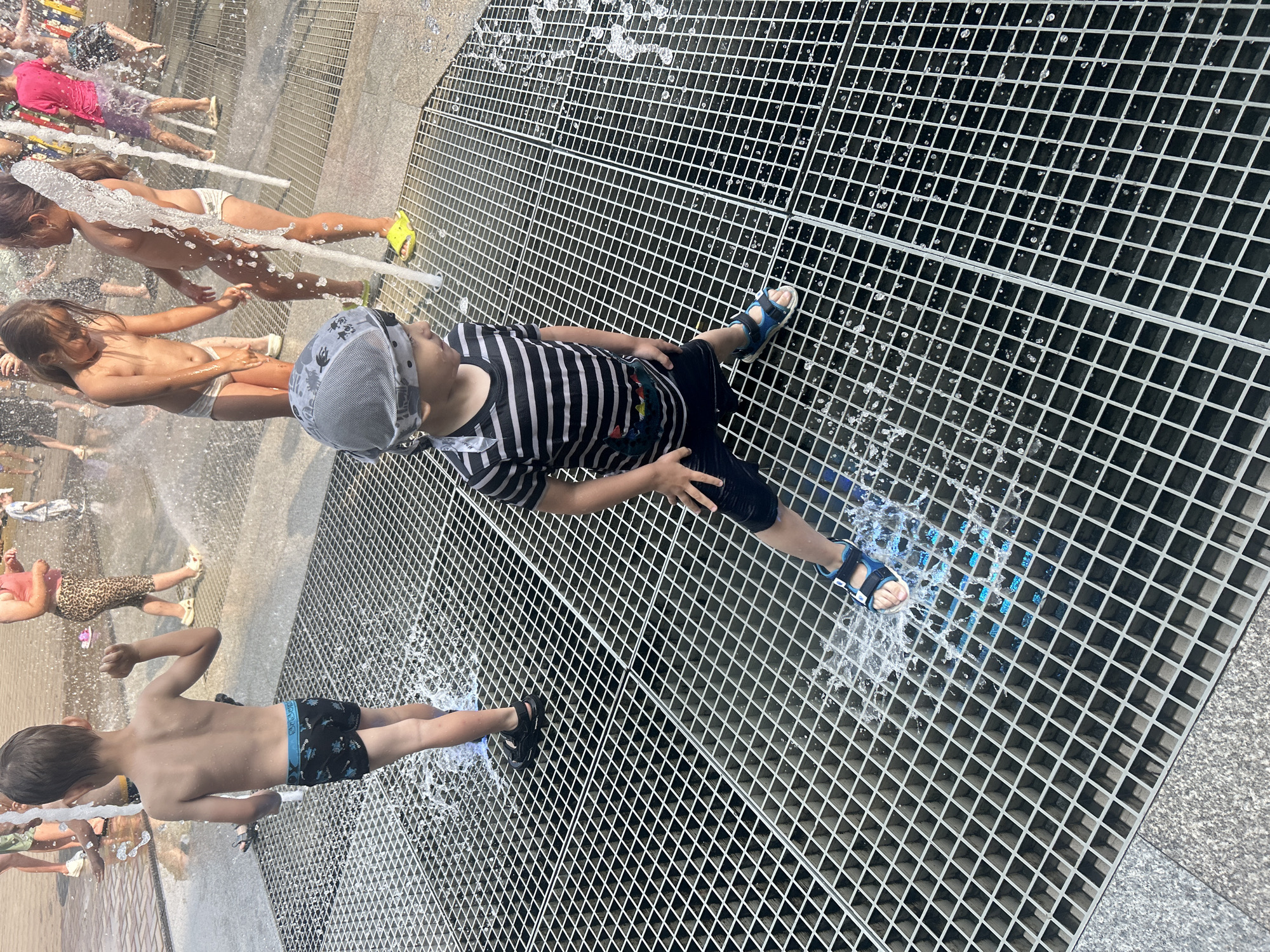 The image depicts a group of children playing in a public splash pad on a sunny day. The setting appears to be an urban area, possibly a park or plaza, with a grate-like surface designed to drain water. Several children are visible, some partially obscured by water jets. One boy, in the foreground and slightly off-center, is wearing a striped t-shirt and dark shorts, with a grey and white patterned cap. He is standing on the grate and appears to be enjoying the water. To his left, another boy in black patterned swim trunks is also being splashed. Further back, a girl in white underwear is visible, as are other individuals and children in the background, some of whom are also getting wet. The water is actively spraying, creating a dynamic and playful atmosphere. The time of day seems to be daytime, given the bright sunlight casting shadows and the overall illumination. The weather is clearly warm enough for children to be playing in water. The overall mood is one of fun and refreshment. No visible text is present in the image.