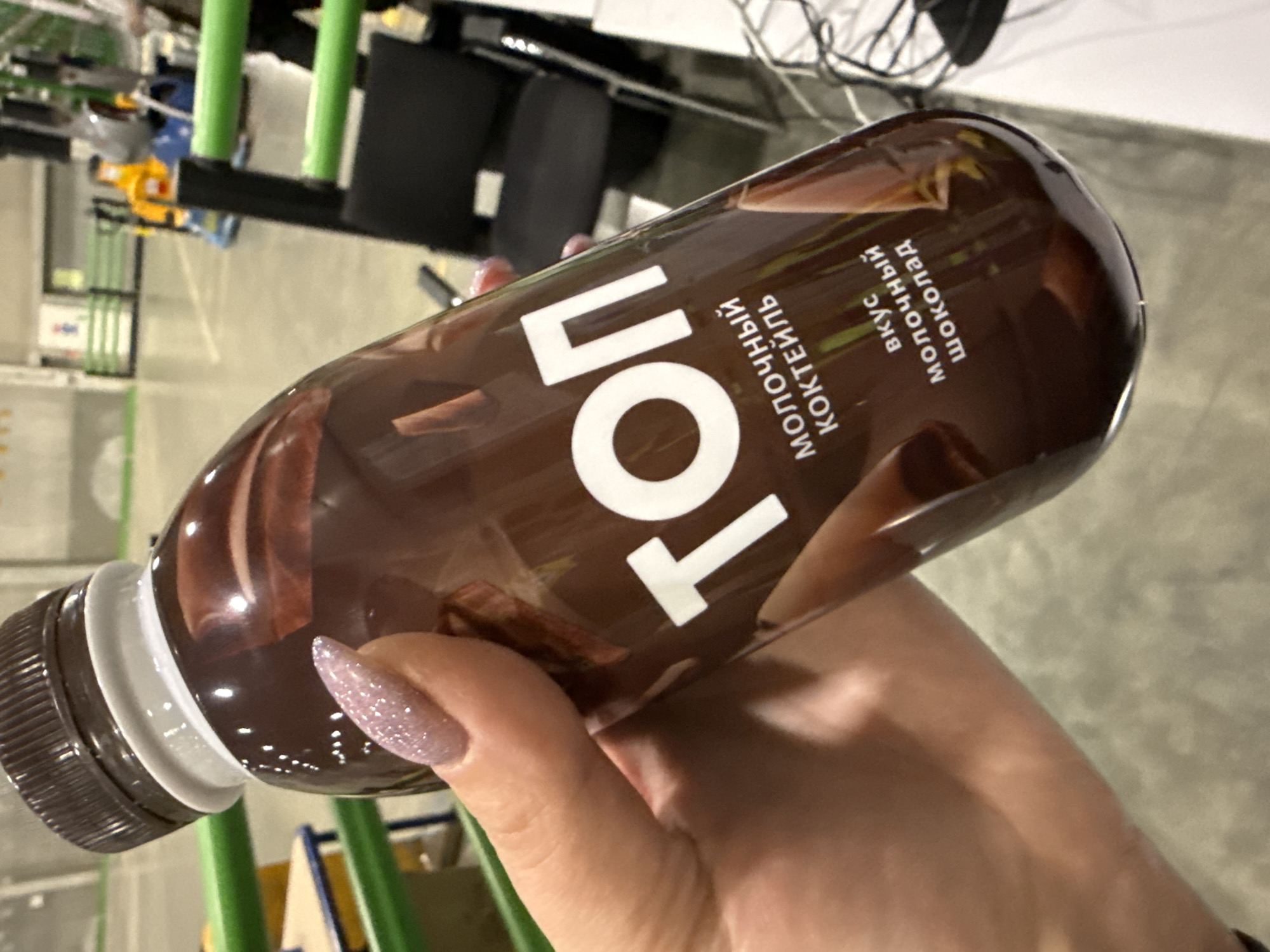 A hand with glittery fingernails is holding a bottle of Top chocolate milk cocktail. The label is in Russian, indicating a chocolate flavor. The background appears to be an indoor setting, possibly an office space or a similar location. Considering the label information, the item likely originates from Russia. It is impossible to confirm the location in the city of Lupanovo, Russia.