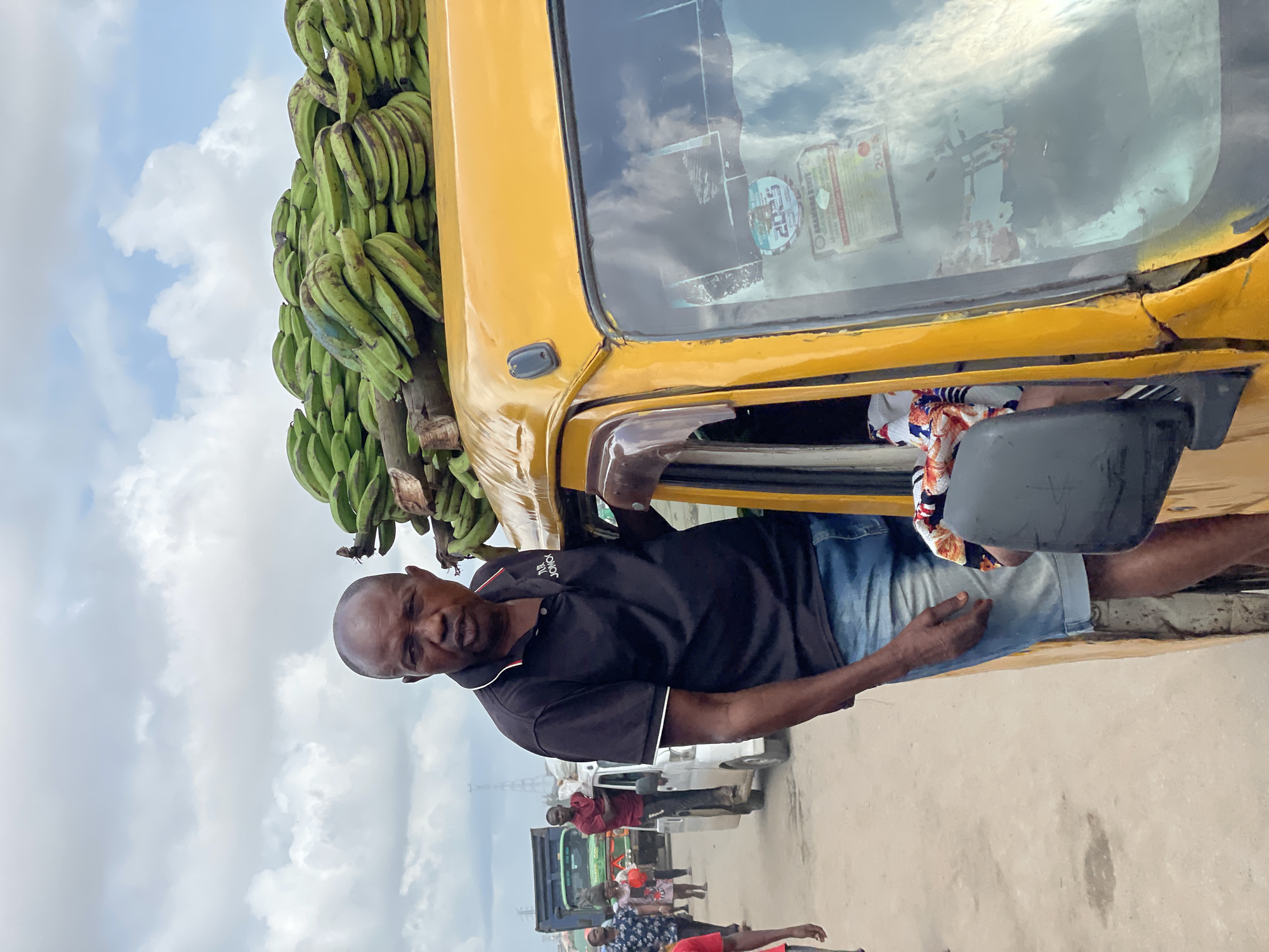 A man stands next to the open door of a yellow motorized tricycle, a large load of green bananas piled high on its roof. He is wearing a black polo shirt with white trim and blue denim shorts. The tricycle appears to be on a street in Fegge, Nigeria, with other vehicles and pedestrians visible in the background. The sky is overcast with some clouds. The man is looking directly at the camera with a neutral expression.