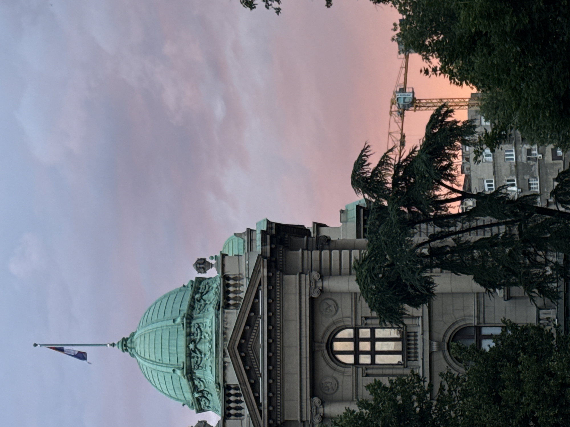 The image shows a view of the National Museum of Serbia in Belgrade, Serbia, at dusk. The museum is a large, imposing building with a green copper dome and classical architectural details, including columns and ornate carvings. A Serbian flag flies from a flagpole on top of the dome. In the foreground, there are dark green trees, and in the background, a construction crane is visible against a pink and orange sky. The sky is filled with soft, diffused clouds, indicating sunset or sunrise. The scene appears to be taken from a slightly elevated position, looking towards the museum and the surrounding urban landscape.