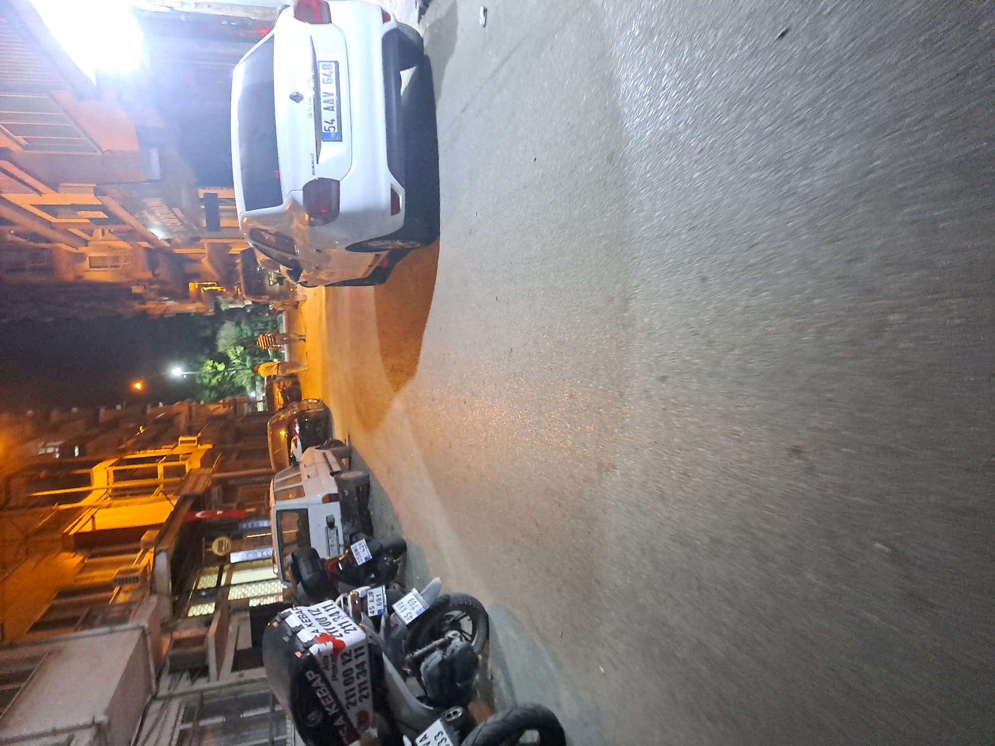 The image depicts a street scene at night in Manisa, Turkey, with several vehicles parked on the side of the road. A white sedan with the license plate "54 AAV 648" is prominently featured, facing away from the camera. To its left, a white utility vehicle is parked. Further to the left, a black scooter is visible with text on its side that includes "A KEBAB" and phone numbers. Another scooter is parked behind it. Two individuals are walking on the sidewalk in the background, illuminated by streetlights. The buildings lining the street are multi-story structures with visible windows and balconies, suggesting a residential or commercial area. The road surface appears wet, reflecting the artificial light sources.