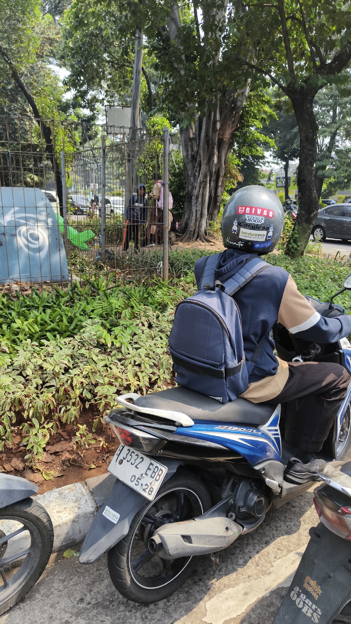 A person wearing a helmet and a backpack is sitting on a blue and white motorcycle. The motorcycle is parked on a paved surface next to a curb with a sidewalk. Behind the motorcycle, there is a fence with some greenery and trees. In the background, there are more trees and a few cars. The scene appears to be a street in a city. The license plate on the motorcycle reads "B 3532 EBB" and has a date stamp "05:28".