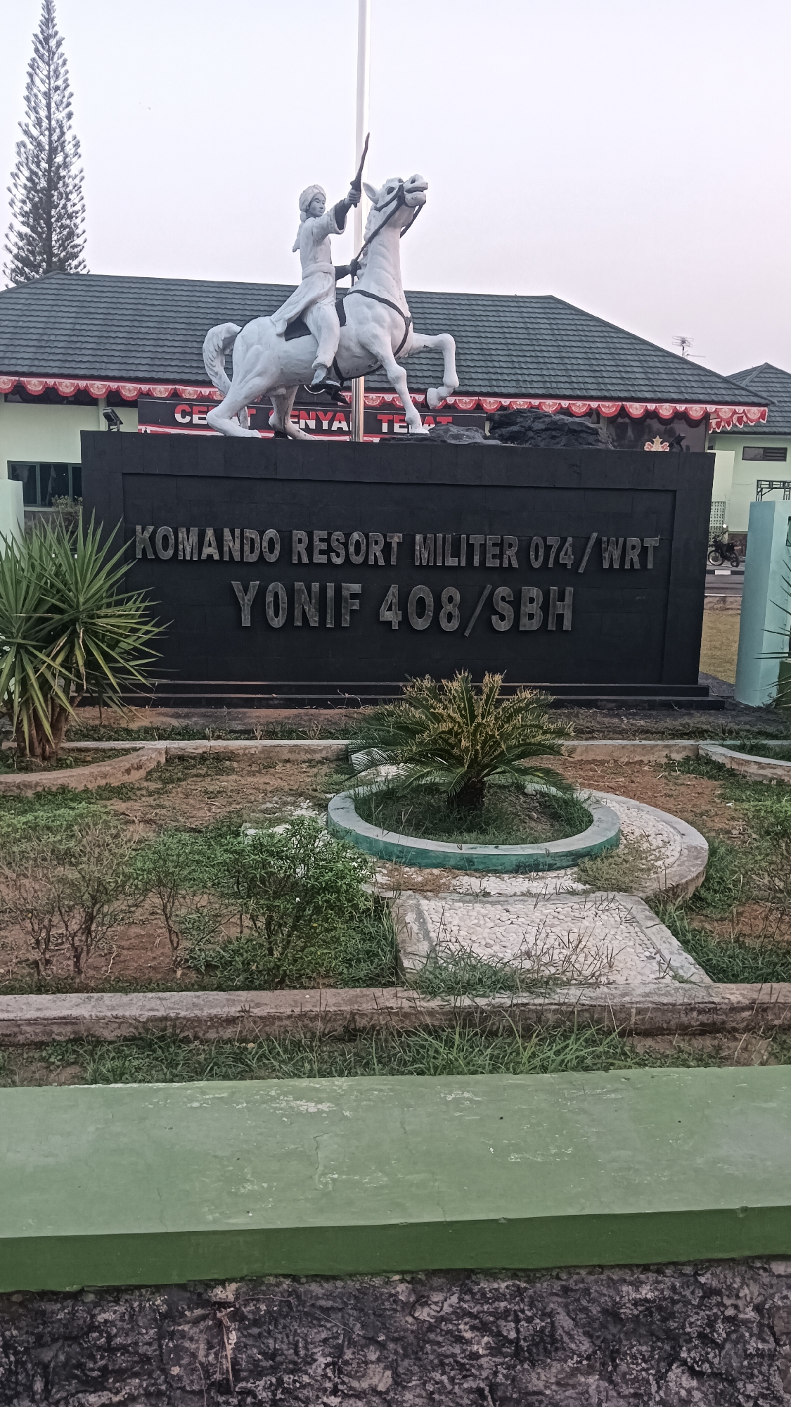 The image shows a statue of a person on horseback in front of a building. The statue is white and the person is wearing traditional clothing. The building is dark and has a large sign that reads "KOMANDO RESORT MILITER 074/WRT YONIF 408/SBH". The building appears to be a military base. The area in front of the building is landscaped with plants and gravel. The sky is overcast. The location is the entrance to Yonif 408/SBH, Sragen, Indonesia.