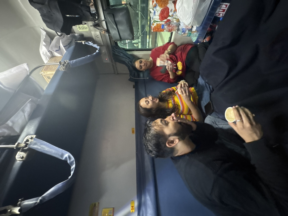Within a train compartment, likely in Asansol, India, three individuals are seated on the lower berth. On the left, a male with dark hair and a beard, wearing a black long-sleeved shirt, holds a white paper cup in his left hand, looking right with his mouth slightly open. To his right, a female with long brown hair and glasses, wearing a striped long-sleeved top and jeans, holds a white paper cup and smiles, looking towards the right. A black mobile phone rests on the blue berth next to her. Further right, a second female with dark hair, wearing a red long-sleeved top with a Mickey Mouse graphic and black pants, holds a clear plastic cup, looking left with a slight smile.
The compartment features blue lower berths and light-colored walls. Berth numbers "54" and "53" are visible on the wall. An upper berth is present, with luggage including wrapped items and a cardboard box. A blue support strap hangs from the upper berth structure. Near the window on the right, power outlets are visible. A black shoulder bag hangs near the window, below which a small table-like surface holds two plastic water bottles, a white plastic bag, and a colorful, possibly floral-patterned item. Through the window, portions of a railway platform and ambient lighting are visible, indicating the train is likely stationary at a station. A dark blue blanket covers parts of the lower berth and the legs of the leftmost individual. All three individuals appear engaged in conversation while holding beverages.