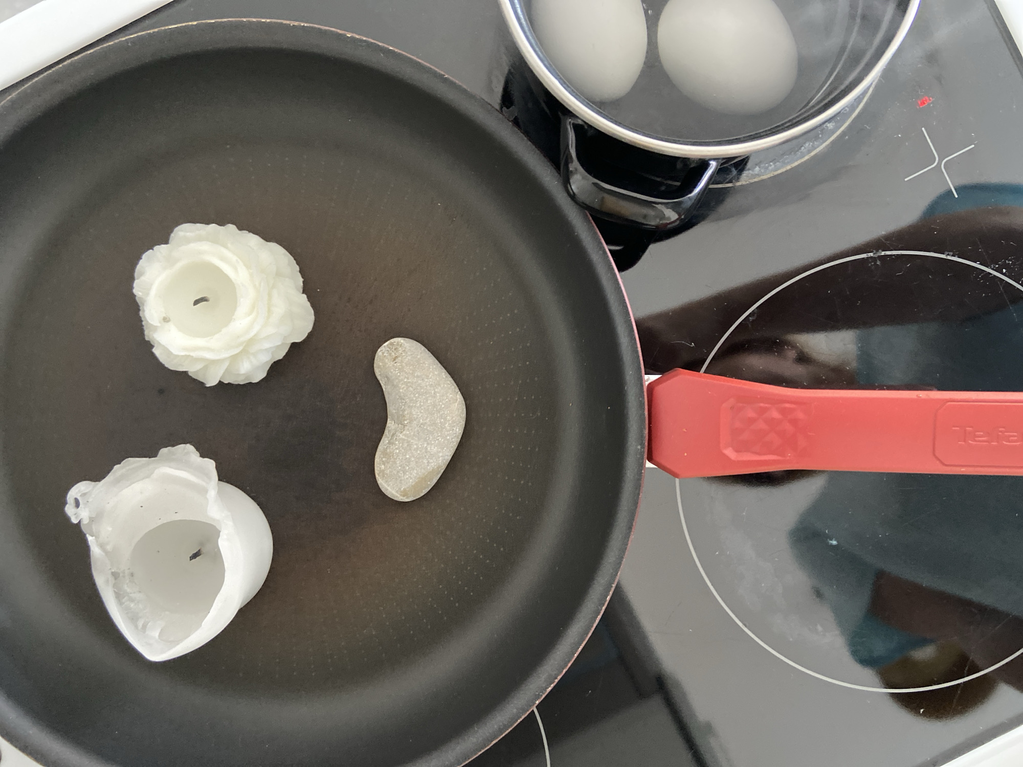 The scene shows a stovetop with a black electric cooktop. A black frying pan with a red handle sits on the cooktop, containing a white, flower-shaped candle, a broken white candle, and a smooth gray stone shaped like a bean. To the upper right, a small pot with two white eggs in water is also on the cooktop, which is turned on and indicated by a red light. A reflection is seen on the cooktop, and a "Tefal" logo is visible on the frying pan's handle. The location in the city of Budva, Montenegro is unknown.