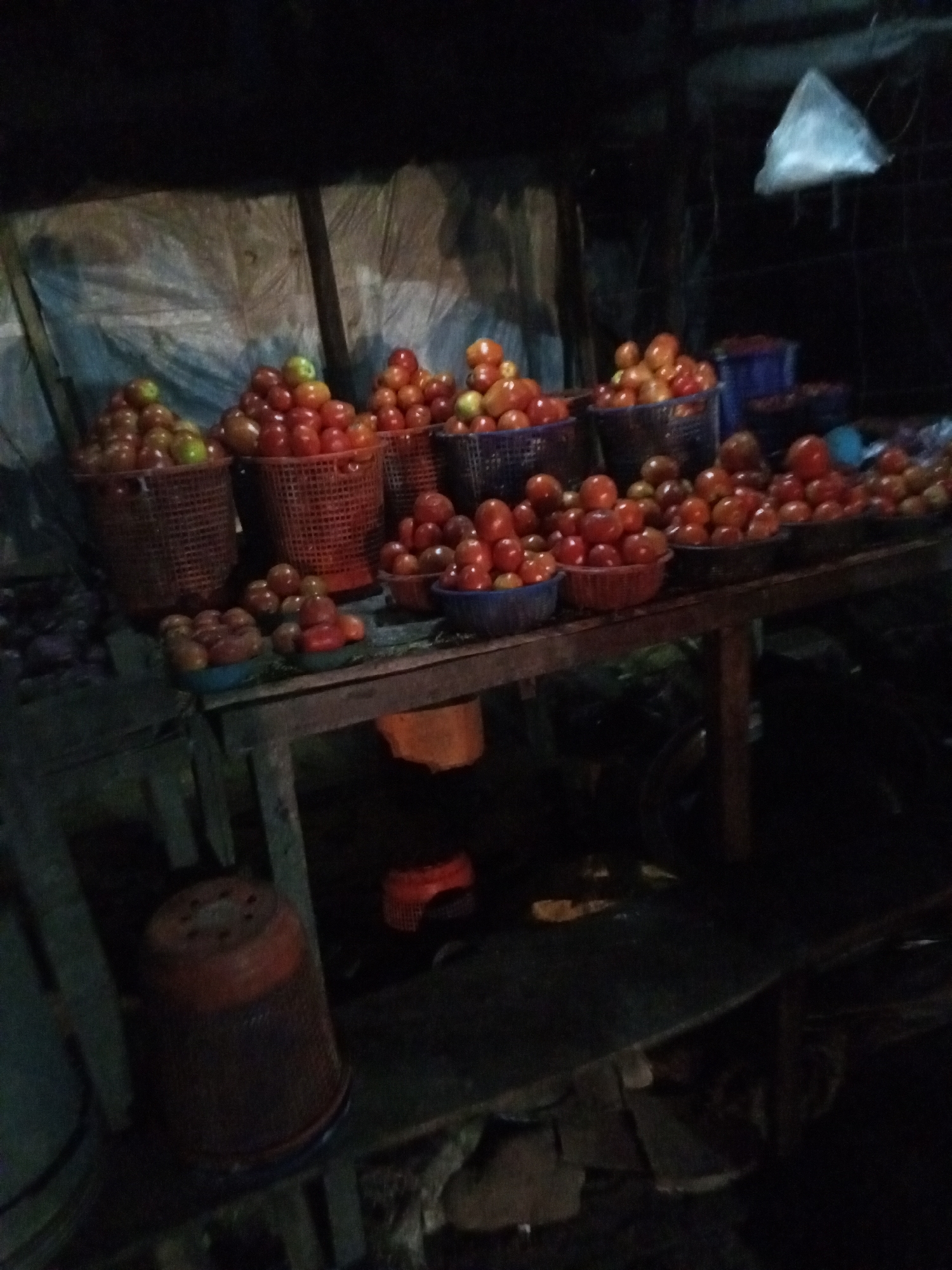 In Lokoja, Nigeria, a dimly lit market stall displays an array of tomatoes. Tomatoes are arranged in woven baskets and plastic bowls atop a weathered wooden table. Additional items sit beneath the table and to the left, including plastic buckets. Above and behind the table is a makeshift shelter. A plastic bag hangs from the roof. Overall, the scene conveys a sense of a small business at night.