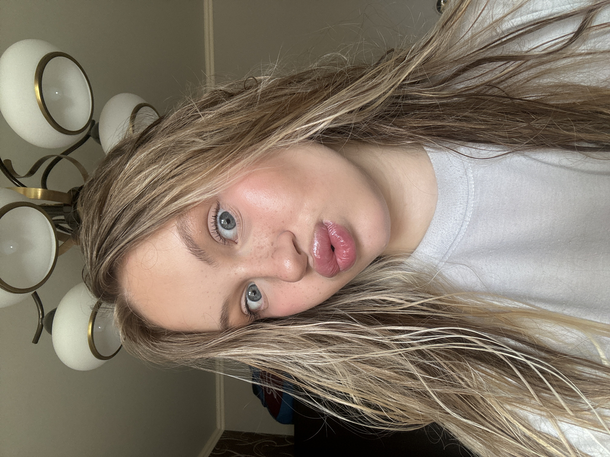 The image shows a close-up, sideways portrait of a young person with blonde hair. The person is wearing a white t-shirt. Their eyes are open and looking towards the camera, with a slight pout to their lips. Freckles are visible on their cheeks. To the left of the person, a portion of a light fixture with three white globes and gold accents is visible. The background is a plain wall, and a glimpse of a patterned floor is seen at the bottom. The lighting suggests it could be daytime, though the interior setting makes it difficult to determine definitively. The overall mood is casual and informal. There is no visible text in the image.