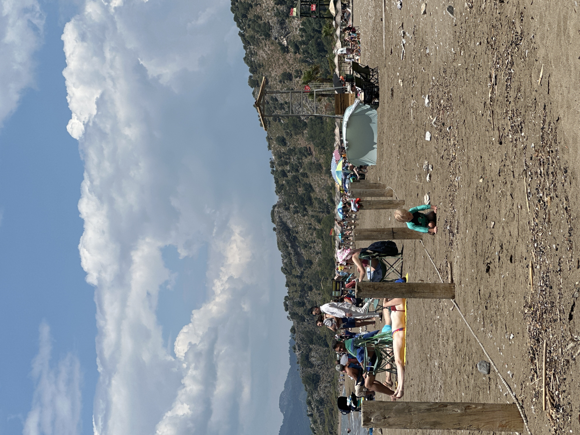 The image shows a beach scene in Ortaca, Turkey, with a variety of individuals and objects visible. The beach is sandy and littered with debris, with people sitting on chairs and blankets. There is a small child playing on the sand. In the background is a mountain covered in vegetation. The sky is blue with white clouds. The scene is marked by wooden posts and ropes, possibly indicating designated areas or boundaries. A lifeguard tower and various beach umbrellas are also visible in the background.
