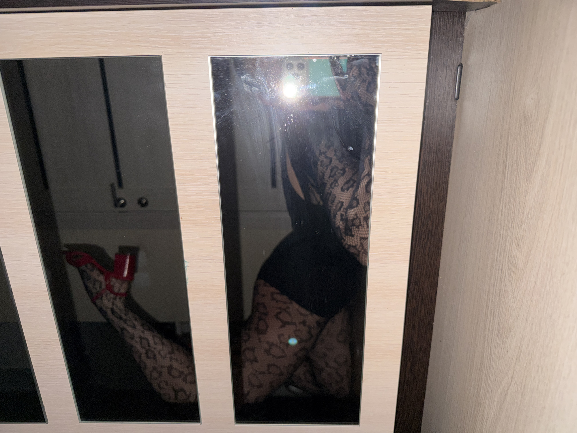 The image captures a reflection of a person in two mirrored cabinet doors, likely part of a wardrobe or storage unit, set indoors in what appears to be a residential space. The cabinet features a light wood-grain finish on its frame, with darker wooden trim along the top, bottom, and between the mirrored panels. To the right, a plain, textured light-colored wall is visible.
The reflected person is wearing a black outfit, possibly a bodysuit or top and shorts, paired with sheer hosiery featuring a distinctive leopard-print or similar animal-print pattern. They are also wearing vibrant red high-heeled shoes. The individual is striking a pose, with one leg visibly bent and the other possibly extended, suggesting a deliberate photo session.
The photograph is a selfie, as evidenced by the bright flash and the visible outline of a smartphone held up in the reflection, near the top center. The phone's camera lenses are also discernible. The mirrors exhibit some dust or smudges. No text is visible in the image. The scene is located in Izhevsk, Russia.