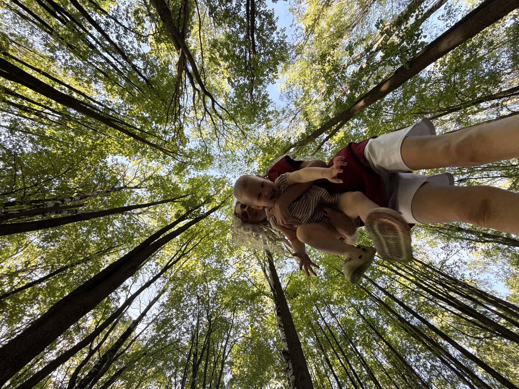 The photograph, taken from a low angle looking upward, depicts a woman holding a baby in a forest. The woman is wearing light-colored shorts, a red shirt, and has long, light-colored hair. She is holding the baby, who is wearing a striped shirt, towards the camera. The forest is dense with tall trees reaching towards the sky, partially obscuring the blue sky above. The leaves on the trees are green. The location is Шебунино, Russia.