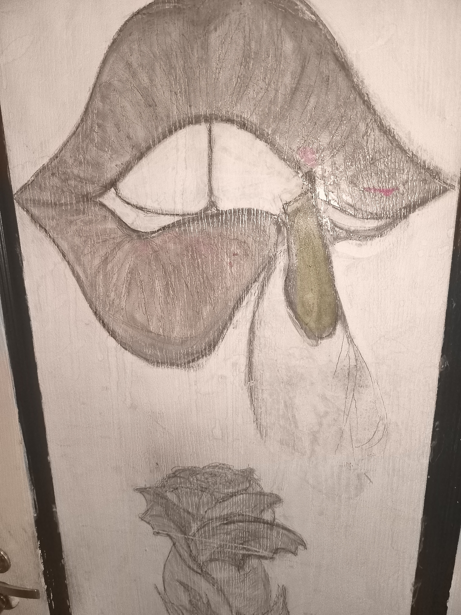 The image is a close-up, slightly angled shot of a painting on a light-colored, textured surface, likely a wall or door. The painting depicts a stylized representation of lips with a teardrop or elongated shape falling from the lower lip. The lips are rendered in shades of gray and brown, with subtle pink accents on the inner edges. The falling element is colored in shades of green and gray. Below the lips, a sketch of a flower, possibly a rose, is visible. The overall aesthetic is artistic and somewhat abstract. The image is not a photograph of a scene in Riga, Latvia, but rather a close-up of a painting. There are no people, actions, or interactions depicted, only the artwork itself. The environment appears to be indoors, given the texture of the surface and the presence of what looks like a door frame on the left edge of the image.