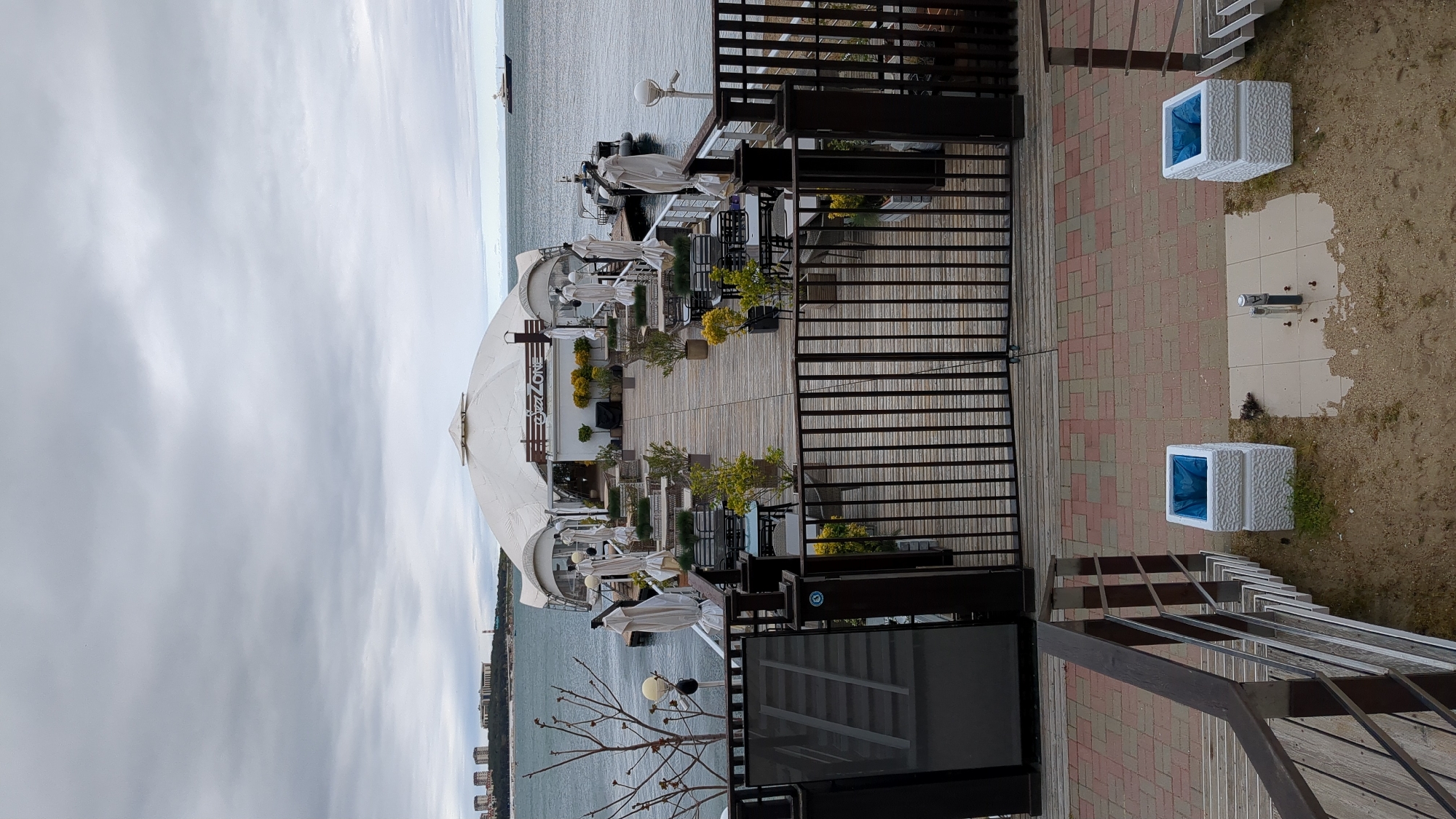 The image depicts a waterfront restaurant called "PizzaZone" in Gelendzhik, Russia. The restaurant has a large white domed structure with "PizzaZone" written on its side. The restaurant's outdoor seating area is visible, featuring tables, chairs, and white umbrellas. The area is decorated with plants in pots. A wooden deck extends from the restaurant. There's a metal railing separating the deck from a brick-paved area. Two white square trash receptacles with blue liners are present near the railing. A metal staircase leads up to a higher level. In the background is the sea, with a cloudy sky above. Buildings are visible along the shoreline.