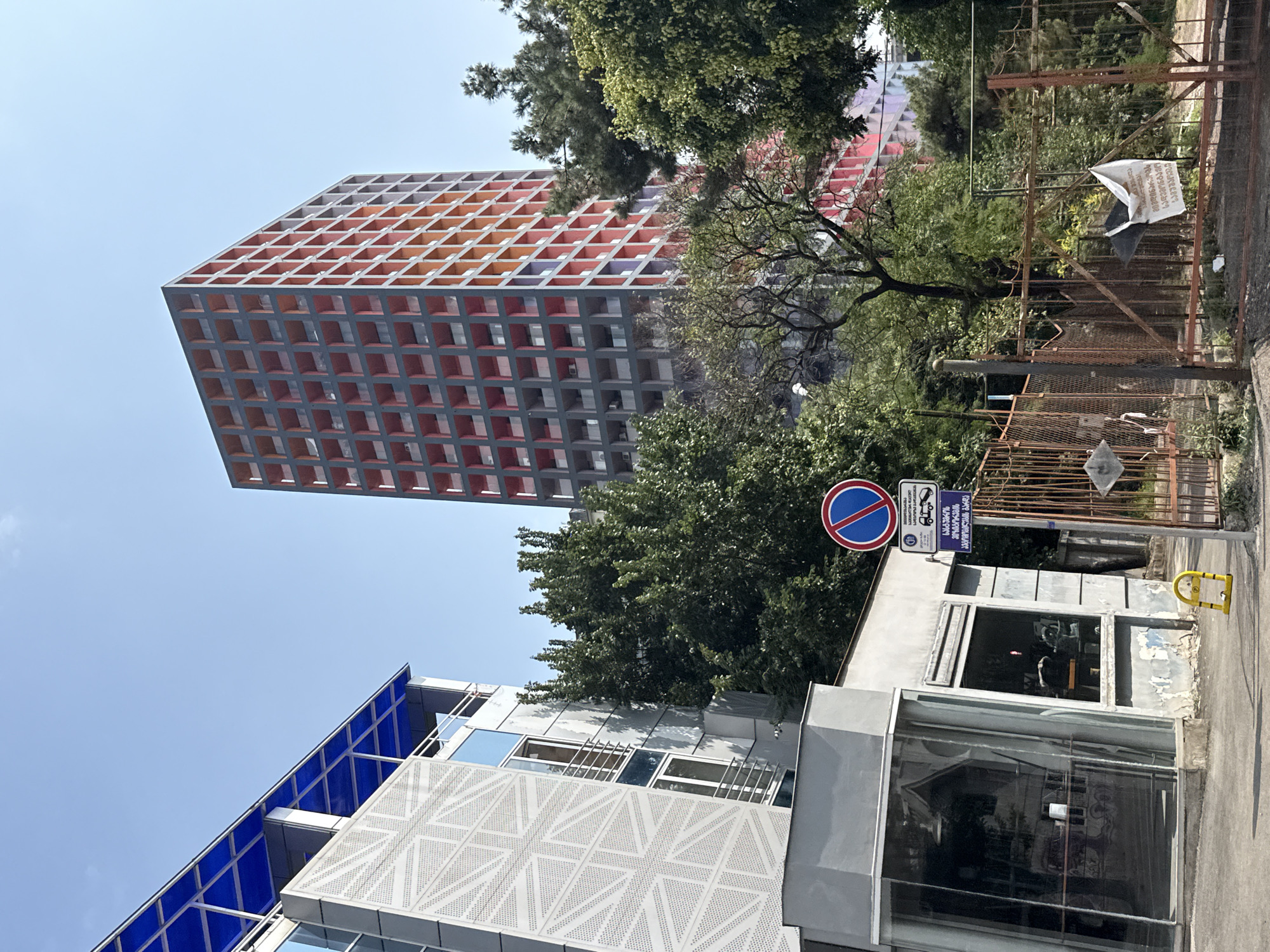 The image depicts an urban street scene under a clear blue sky. On the left, a modern building with a distinctive geometric facade and a blue canopy stands. To the right of it, a contemporary high-rise building with a grid of red and orange balconies dominates the background. Lush green trees partially obscure the view of the buildings. In the foreground, a weathered fence encloses an area, and a street sign with international symbols and Georgian script is visible. A yellow parking bollard is situated on the pavement. The scene appears to be in a city with a mix of modern and older architecture, consistent with an urban environment like Tbilisi, Georgia.