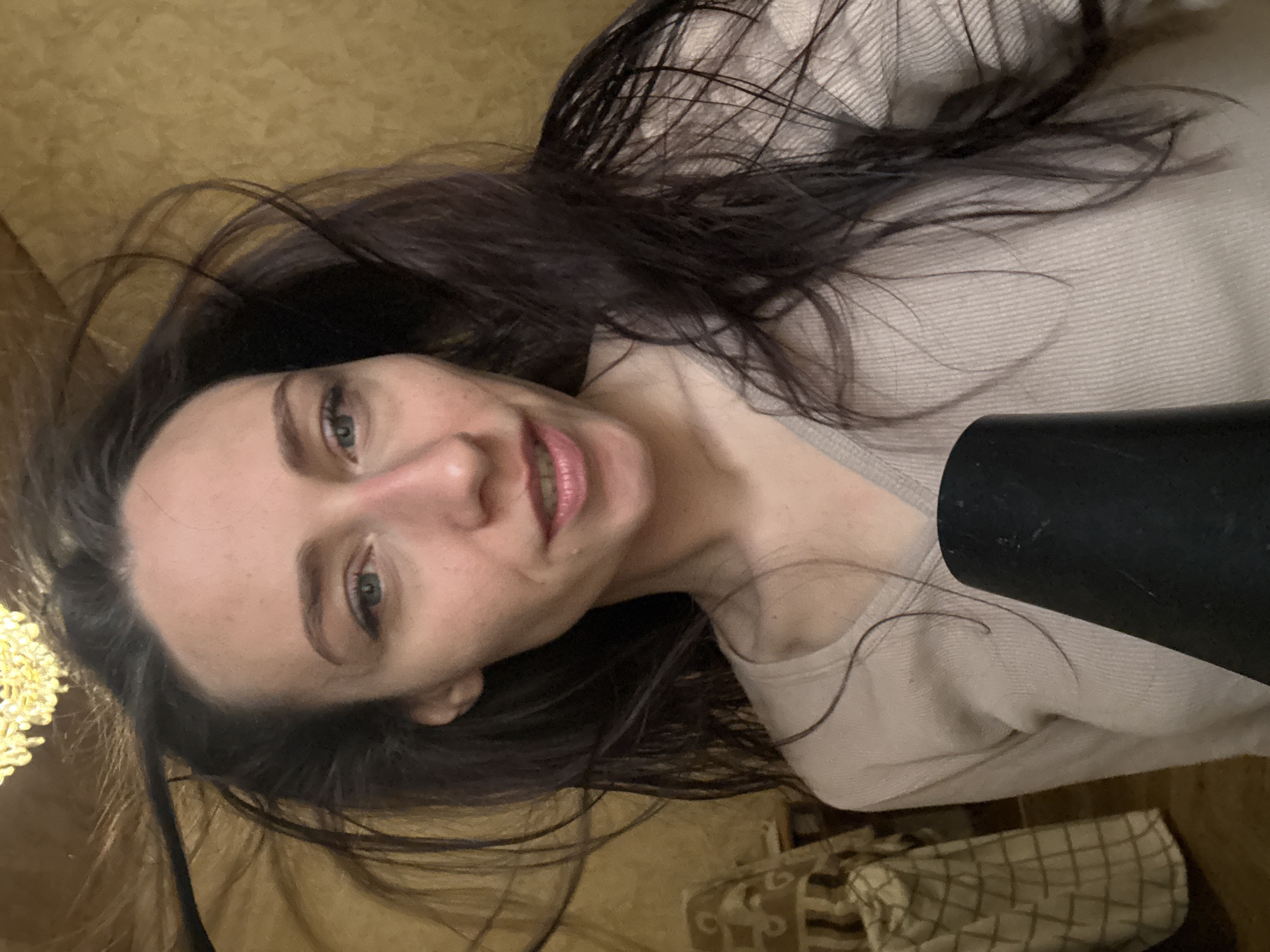 A woman with long, dark hair is lying on a surface, possibly a bed or floor. She is wearing a light-colored sweater. Part of a black object, possibly a hairdryer, is visible in the lower right of the frame. In the background, there is what appears to be a light fixture and patterned fabric. The location in Saint Petersburg, Russia, is not discernible from the image.
