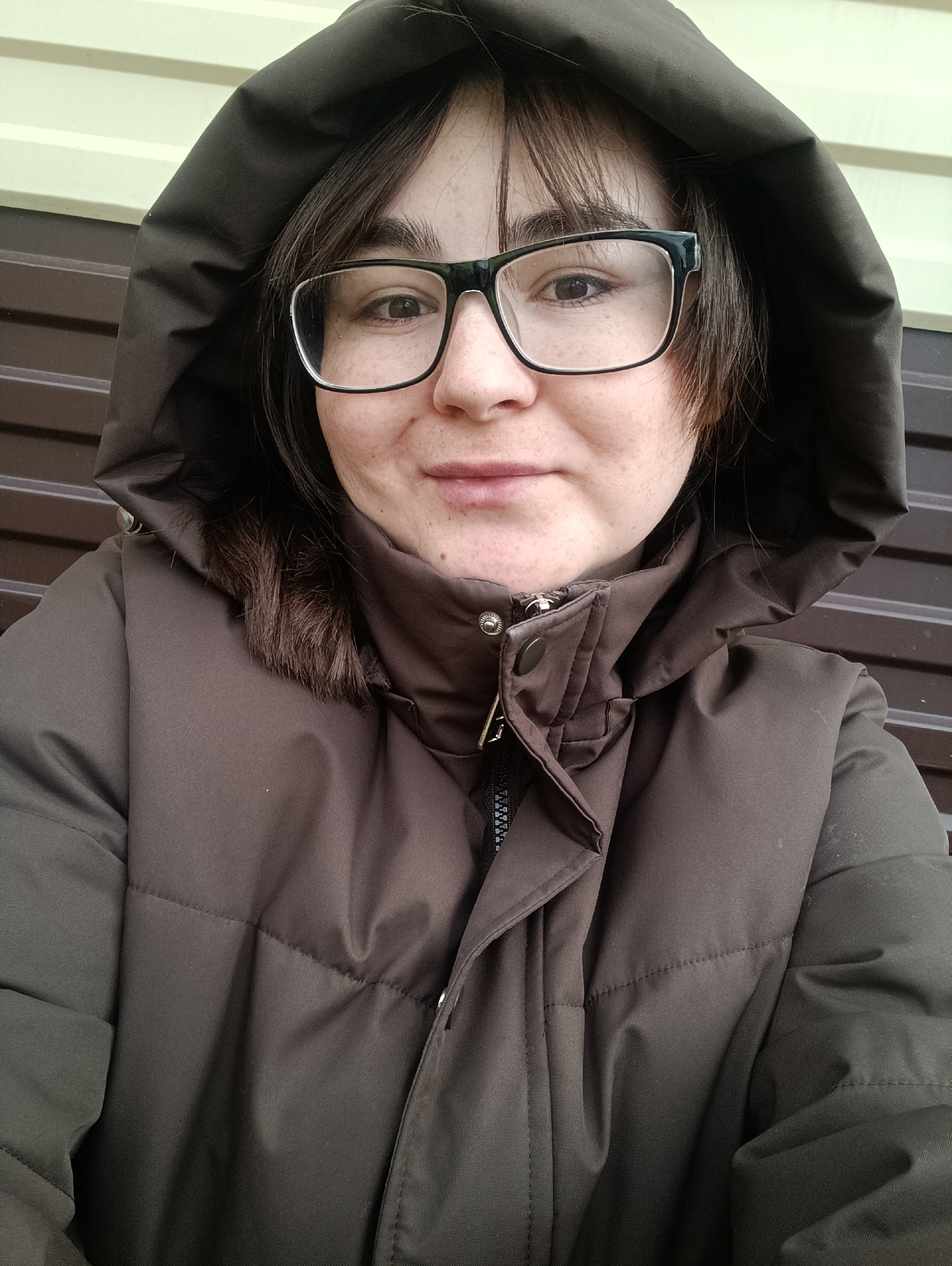 A woman with brown hair and glasses wears a brown hooded puffer jacket. She is facing the camera with a neutral expression. The background consists of light yellow and brown horizontal siding. The location is unknown, but the city name of Barda, Russia has been specified, and the description is limited to the visual content of the photograph.