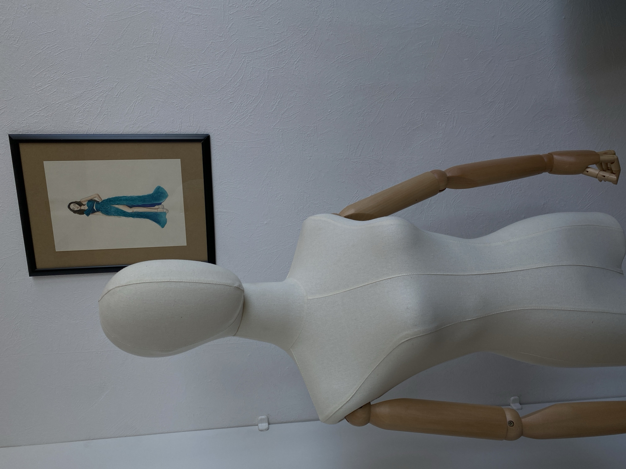 A tan and white female mannequin is shown in front of a black-framed picture on a white wall. The mannequin has a fabric-covered head and torso with wooden arms. The picture is a drawing of a woman in a flowing blue dress. The background is a textured white wall.
The location in the city of Izhevsk, Russia is not discernible from the image.