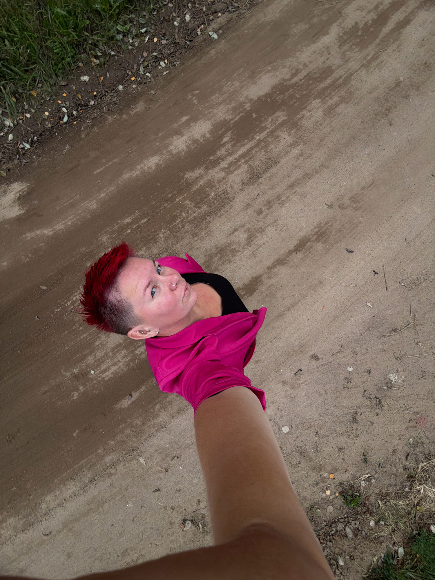 A woman with bright red, spiky hair and blue eyes is looking upwards towards the camera, which is held at a high angle, creating a worm's-eye view. She is wearing a black top and a bright pink hooded jacket, with the hood partially obscuring her hair. She has earrings and a nose ring. She appears to be standing on a dirt path next to a grassy area. The path is light brown and appears somewhat worn, with some lighter streaks indicating moisture or different soil composition. Scattered leaves and debris are visible on the path and in the grass. The overall setting suggests an outdoor environment, possibly a park or a rural road within the city of Chaikovsky, Russia.
