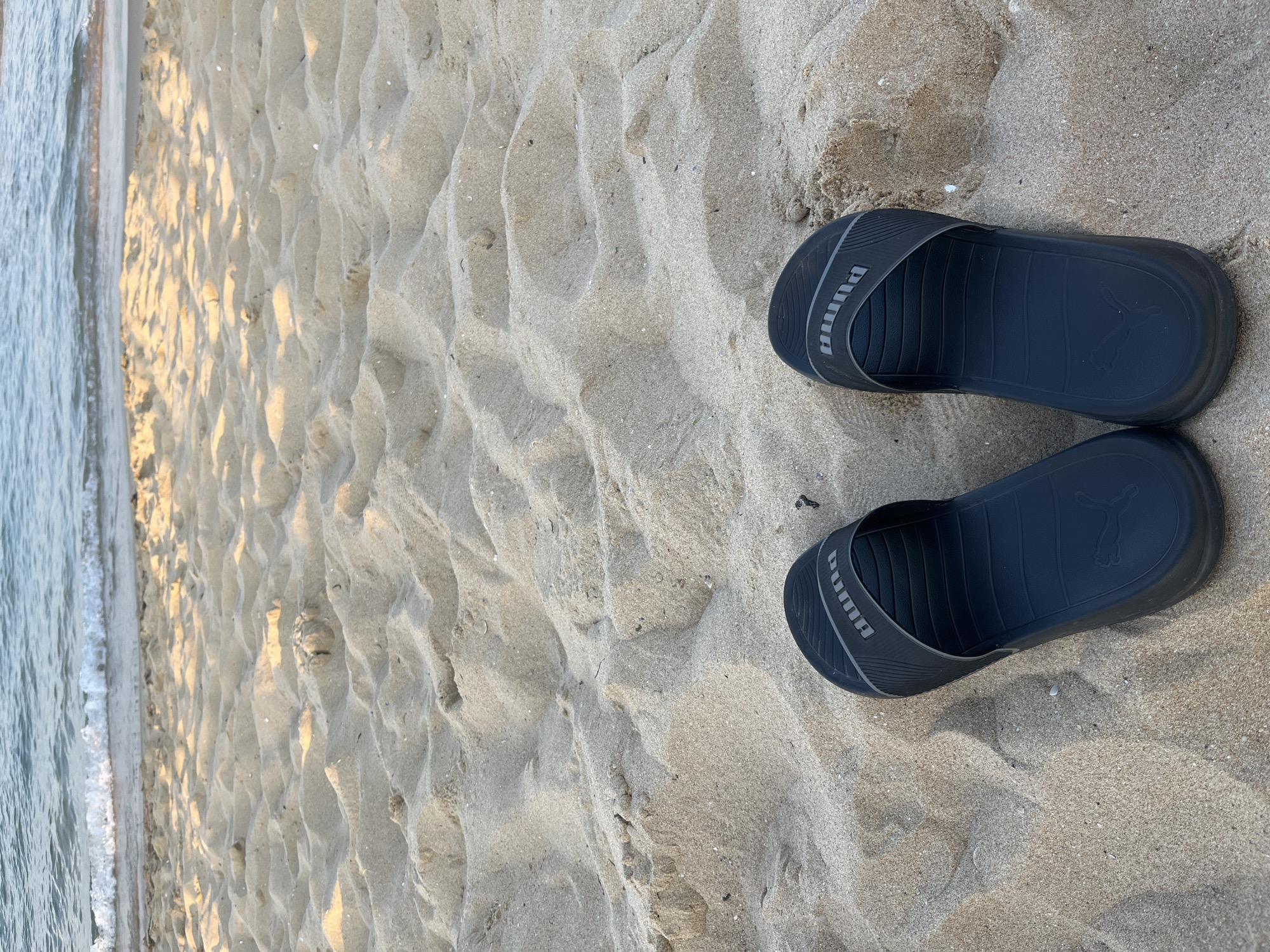 A pair of black Puma slide sandals are positioned on a sandy beach, with the ocean's gentle waves visible in the background. The sand is light brown and has uneven textures, suggesting it is a natural beach. The sandals are facing the viewer, and both have the Puma logo visible on the strap and the footbed. The scene appears to be an outdoor, daytime setting on a beach in Nesebar, Bulgaria. There are no people or other objects in the frame, besides the sandals, sand, and sea.