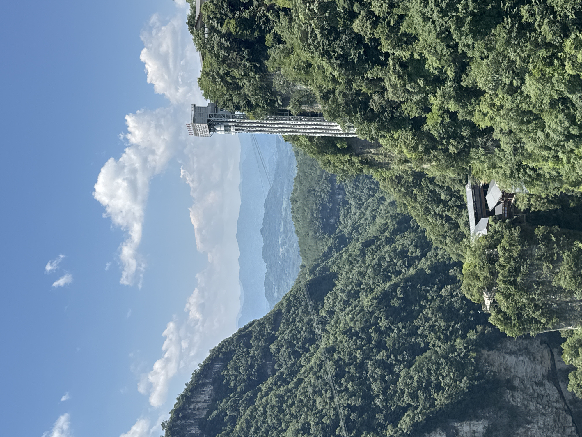 This image captures a dramatic view of a deep canyon in Zhangjiajie, China, dominated by lush green vegetation covering steep, rocky cliffs. A towering outdoor elevator, constructed with a white metal frame, is prominently featured on the right side of the canyon, ascending alongside the cliff face. The elevator's structure is detailed and includes glass-enclosed cabins visible within its framework.
At the base of the canyon, nestled amongst the trees, are traditional-looking buildings with dark tiled roofs, likely part of a tourist facility or village. A wooden walkway or viewing platform can be seen extending from these buildings over the edge of the cliff.
In the background, a series of hazy, blue-hued mountains recede into the distance, suggesting a vast and expansive landscape. The sky is a bright blue, dotted with scattered white cumulus clouds, indicating a clear and sunny day. The overall impression is one of a natural wonder enhanced by human engineering, offering visitors access to breathtaking vistas.