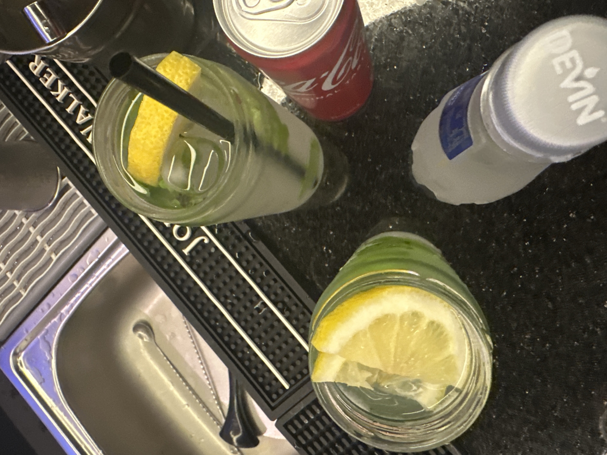 This is a top-down, close-up shot of two drinks, a can of Coca-Cola, and a bottle of Devin water, all placed on a dark, speckled countertop. The scene appears to be set at a bar or a kitchen counter, possibly a domestic setting given the presence of a sink visible on the left.
The two drinks are in clear glasses and contain ice, mint leaves, and slices of lemon. One glass is more prominent in the foreground, with a thick slice of lemon visible at the top, while the other is in the middle ground, with a slice of lemon and a black straw. The Coca-Cola can is red with white lettering, and the Devin water bottle is white with a blue label and a white cap. The countertop is dark and appears wet in places.
The lighting suggests an indoor setting, potentially during the day, given the brightness reflecting off the stainless steel sink. There are no people visible in the image, but the presence of drinks suggests a social gathering or a moment of refreshment. The overall mood is casual and relaxed.
Visible text includes "Coca-Cola" on the can and "DEVIN" on the water bottle. The word "WALKER" is visible on a black bar mat in the background, suggesting a brand or a location.