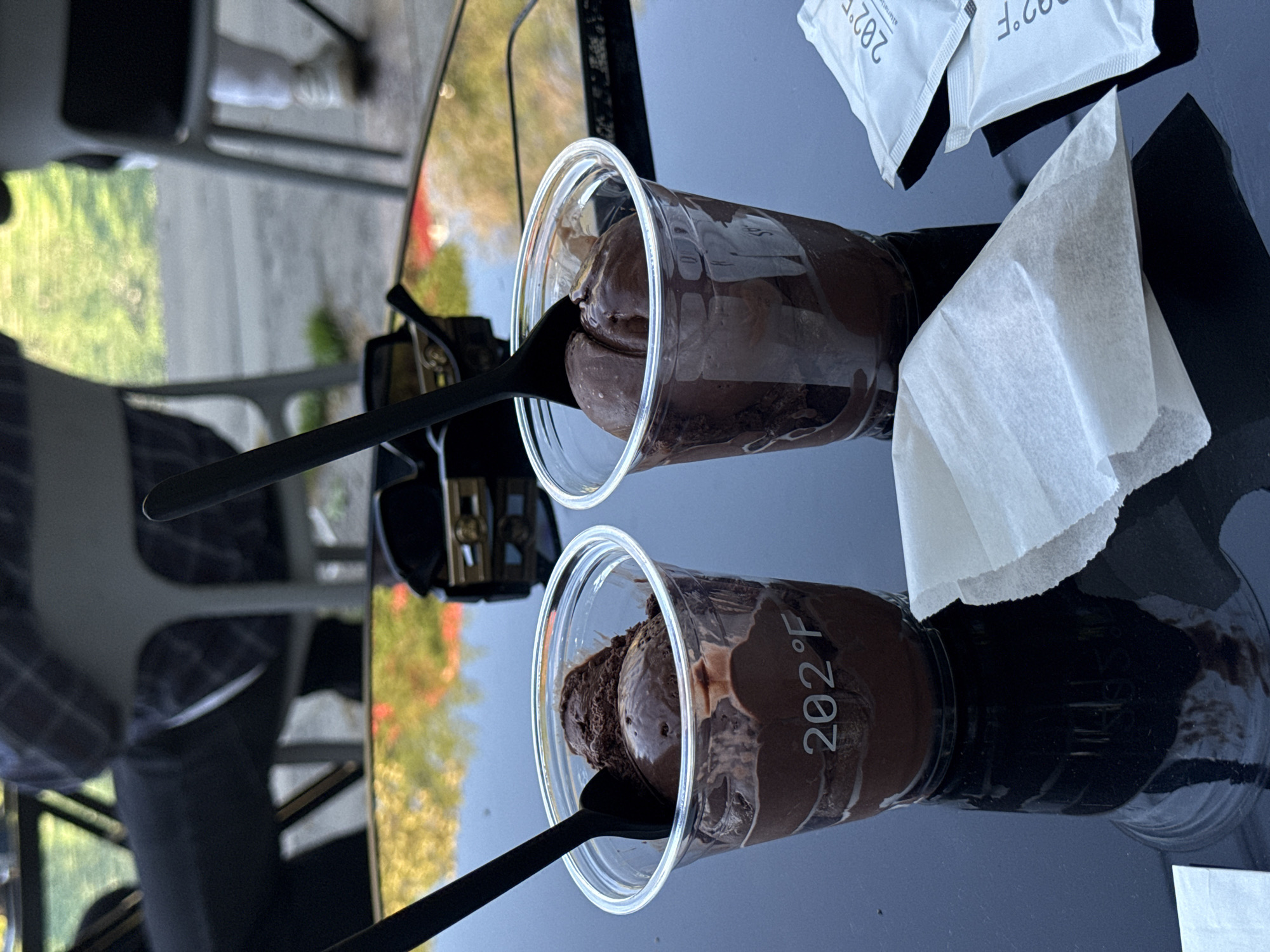 The image shows two clear plastic cups filled with chocolate dessert, likely ice cream or mousse. Each cup has a black spoon inserted into it. Next to the cups, there are two white napkins and two small, white, rectangular packets labeled "202°F." The setting appears to be outdoors, possibly at a cafe or restaurant, with the scene reflected in a glossy black tabletop. The background reveals a blurred glimpse of trees and a building, suggesting an urban environment. Given the "202°F" branding and the visible reflections of the city outside, the scene is highly likely to be at a 202°F cafe location in Yerevan, Armenia. A chair and what appears to be a person's leg are also visible in the corner of the image.