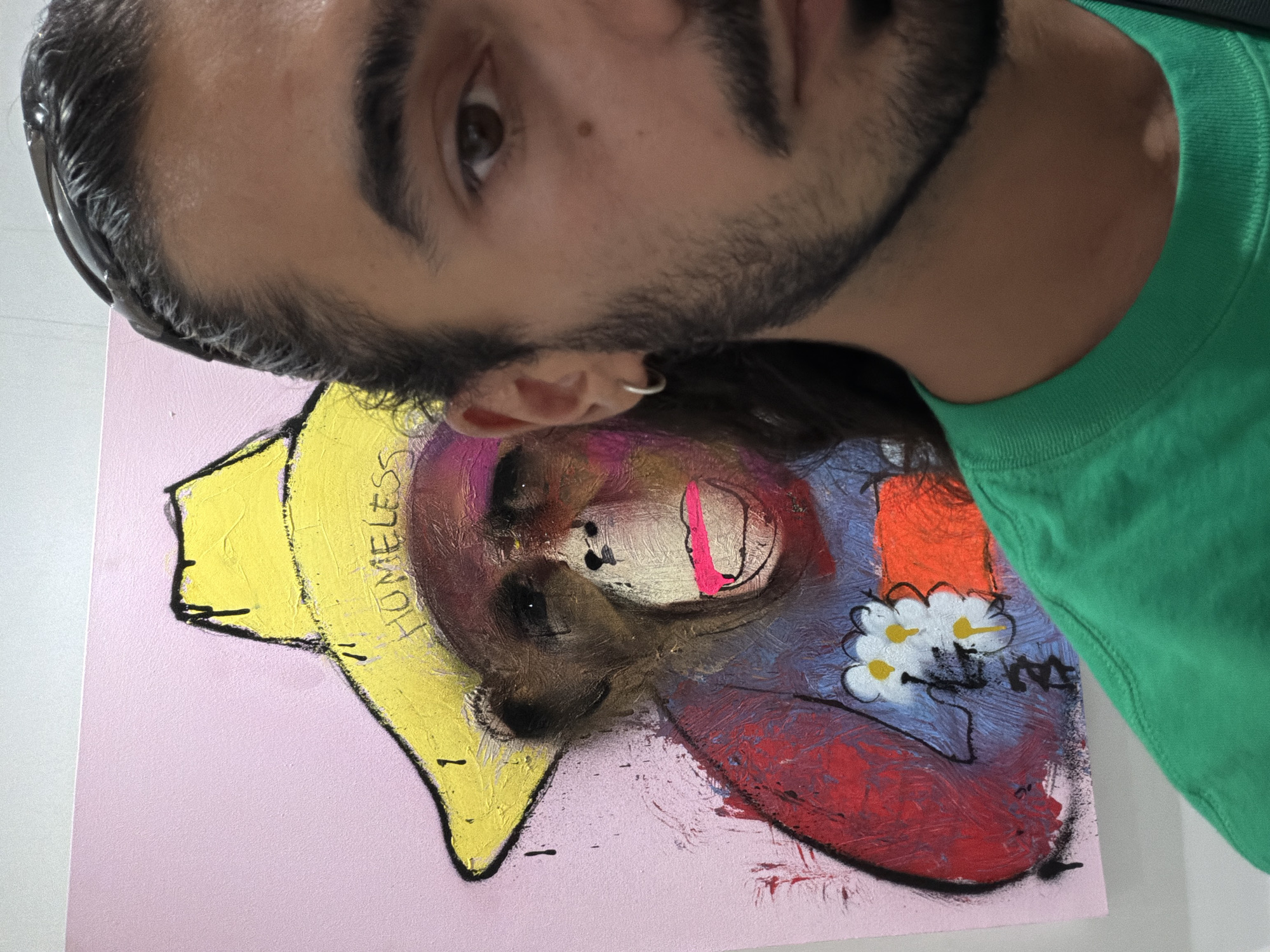 The media file shows a close-up, upside-down, and slightly angled shot of a person's face and upper body, superimposed over a painting. The person has dark hair, a beard, and is wearing a green t-shirt. They are also wearing sunglasses on their head and have a silver hoop earring in their visible ear.
The painting, which occupies the lower portion of the frame, is a colorful abstract depiction of what appears to be a primate wearing a yellow, wide-brimmed hat. The background of the painting is a light pink. The primate has a distressed appearance with bold brushstrokes in shades of purple, red, and brown. There are elements of pink and black used to define facial features, including a bright pink line suggesting a mouth. A small cluster of white flowers with yellow centers is depicted on the primate's chest.
The word "HOMELESS" is visible in black, slightly smudged lettering on the yellow hat in the painting. The overall impression of the image is artistic and perhaps a commentary on social issues, given the visible text. The lighting suggests an indoor setting, and there are no cues about time of day or weather. The focus is on the interplay between the person and the artwork. The location context is Kuta, Indonesia, but there are no specific visual indicators within the image to confirm this.