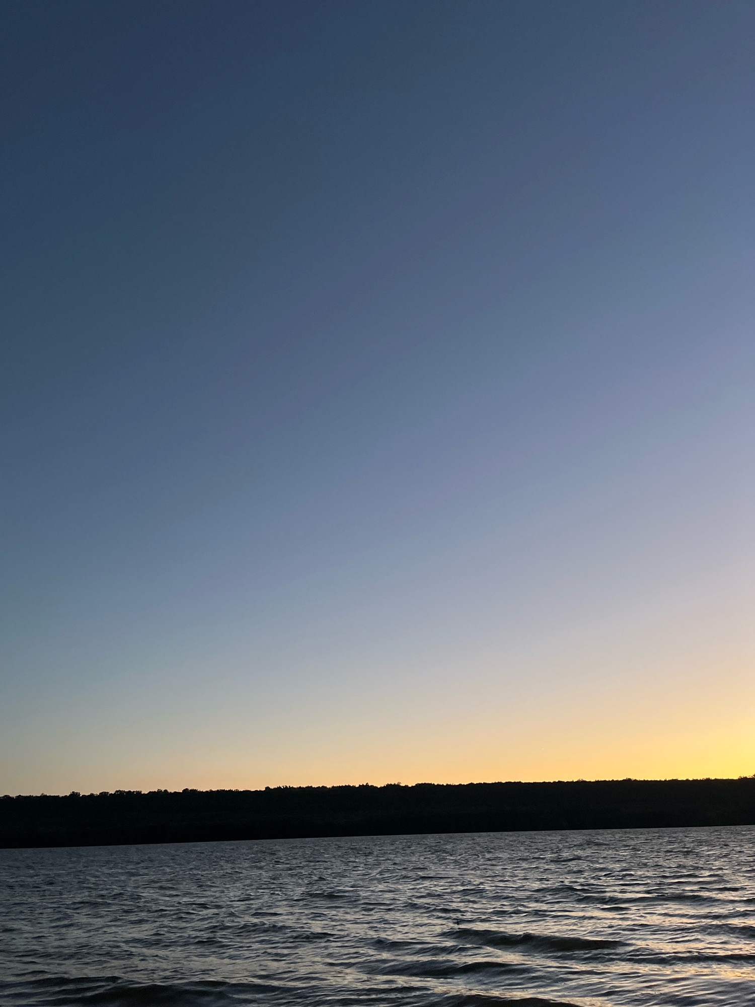 The image depicts a body of water with gentle ripples, likely a lake or river, under a gradient sky that transitions from a deep blue at the top to a soft yellow or orange near the horizon. A silhouetted treeline or dense vegetation forms a dark band along the horizon, separating the water from the sky. There are no people, animals, or man-made structures visible in the scene. The environment appears natural and tranquil. The location in Korzhevo, Moldova, suggests this is a scenic natural landscape within or near the town.