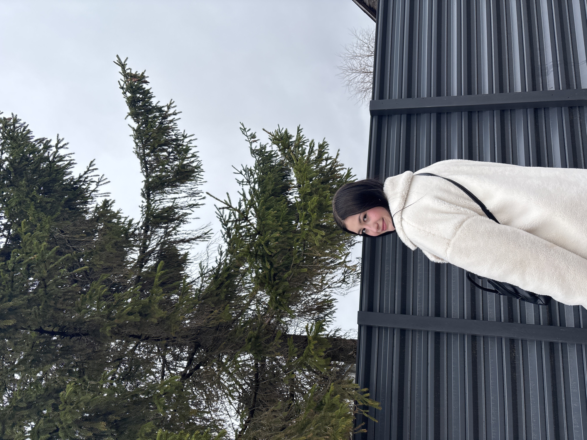 A low-angle shot shows a dark-haired woman wearing a white, fluffy coat standing adjacent to a grey metal building with vertical lines. Part of a tree with green foliage is visible to the left, with a grey sky in the background. The location is Фоки, Russia.