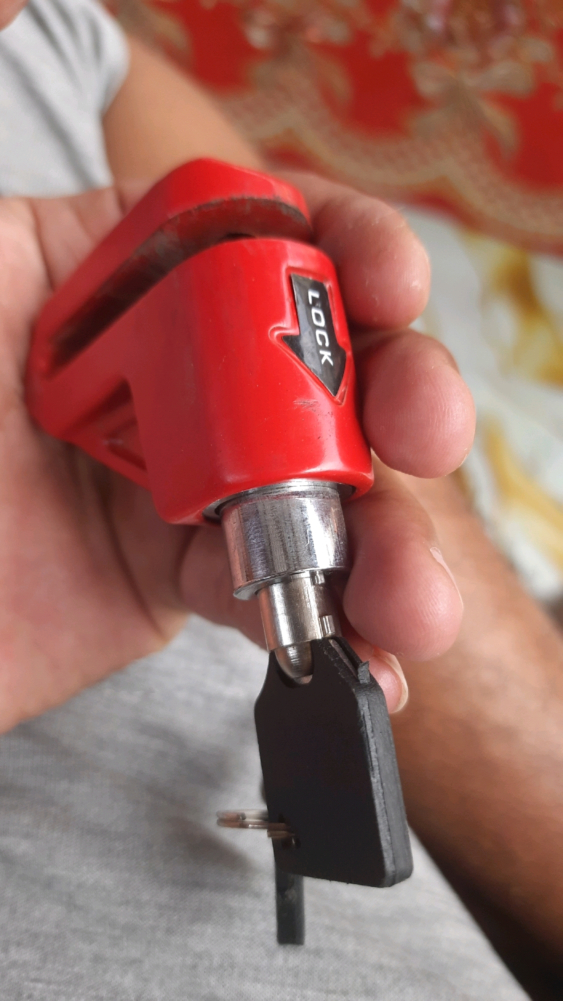 In Kebondalem, Indonesia, a close-up shot captures a person's hand holding a bright red disc lock, a security device typically used for motorcycles or bicycles. The lock has a black arrow pointing to the word "LOCK" inscribed on its side. A black key is inserted into the lock's mechanism, suggesting it is either being locked or unlocked. The background is out of focus but appears to be an indoor setting with patterned fabric, possibly upholstery or curtains. The lighting is natural and appears to be coming from the side. The image focuses on the detail of the lock and key, with the person's skin and clothing visible in the periphery.