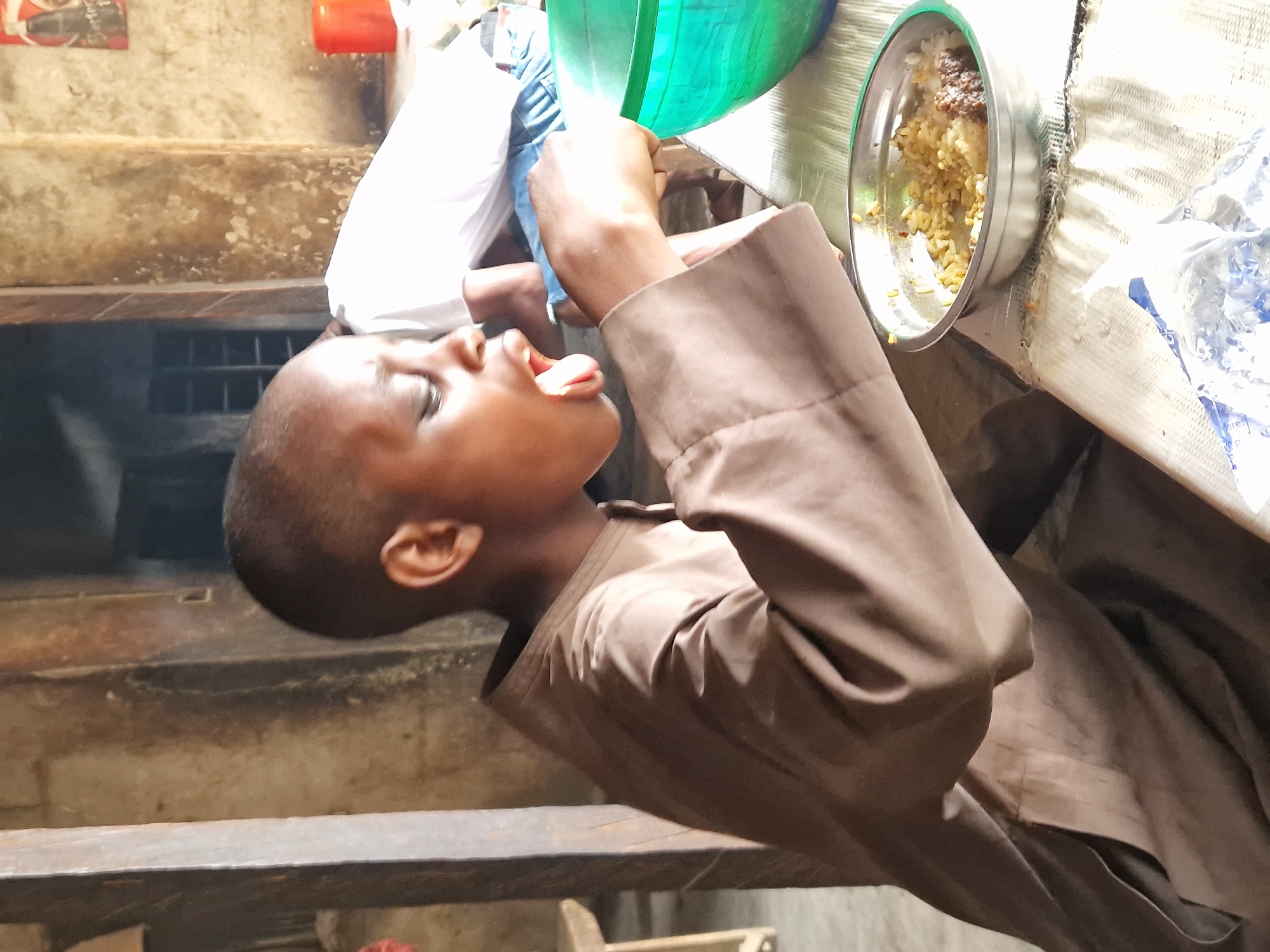 A young boy with a shaved head and a brown kaftan is in the process of eating rice from a metal bowl. His mouth is open, and his tongue is visible as he brings food to it with his hand. He is seated at a table covered with a mat. Beside him is a large green bowl. The background shows a rough, textured wall, a doorway, and a hint of what appears to be a poster or sign on the wall. The scene suggests a simple dining setting, possibly in a home or community space, located in the city of Jos, Nigeria.