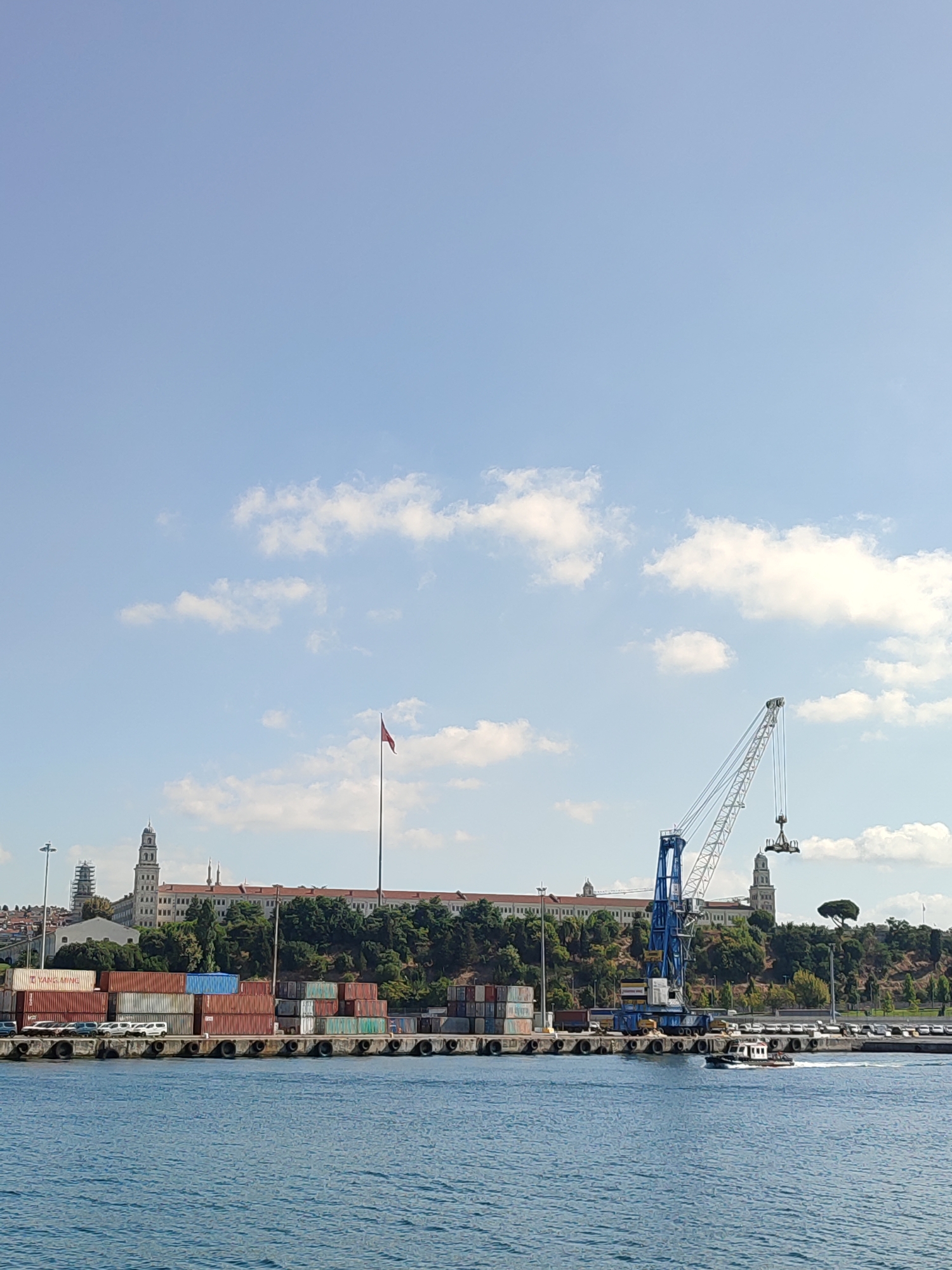 This image shows a waterfront scene in Istanbul, Turkey, with a prominent port facility in the foreground and a large building on a hill in the background. The water is calm and blue, with a small tugboat visible in the middle right of the frame.
The port area is filled with stacked shipping containers, and a large blue and white crane is actively working, lifting a container. Several cars are parked along the quay. Behind the port, a long, multi-story building with many windows stretches across the hill, topped by two distinctive towers. A tall flagpole with a red flag is positioned in front of the building. The hill is covered with lush green trees. The sky is clear blue with scattered white clouds. The overall impression is of a busy, functional port operating alongside historical architecture.