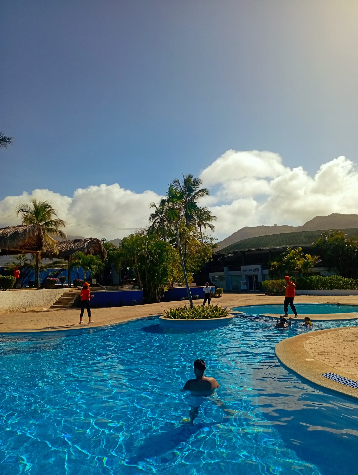 The image depicts a sunny day at a pool complex in Playa El Agua, Venezuela. The complex has multiple interconnected pools with bright blue water. Several people are visible in and around the pools. One man is standing in the foreground pool, facing away from the camera. Other individuals are swimming in an adjacent pool. Several people wearing orange shirts and black pants are positioned around the pool area, some standing at the edges and others walking. Lush tropical vegetation and palm trees surround the pool complex. In the background, there is a building with the words "HESPERIA SPA" visible, suggesting the presence of spa facilities. Mountains are visible further in the background under a blue sky with scattered clouds. There are also thatched-roof cabanas near one side of the pool area. The overall scene portrays a relaxing, tropical vacation setting.