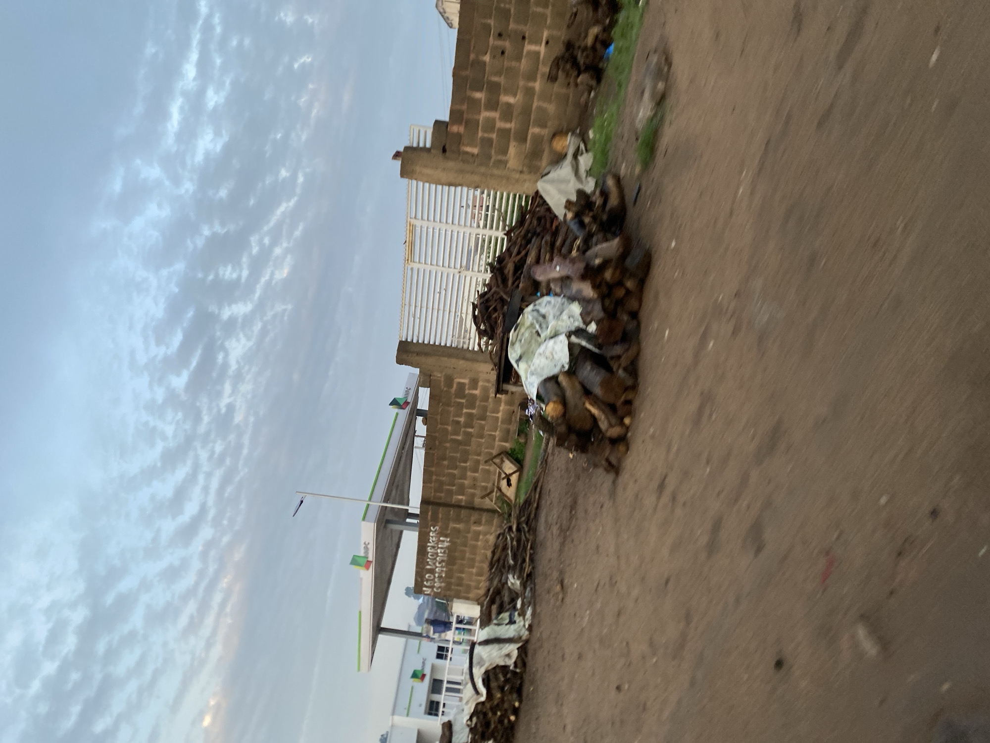 The image shows a dirt road with piles of chopped wood in the foreground. In the background, there is a brick wall and a white metal gate. Behind the wall, there is a gas station with a white canopy and the letters "NNPC" visible. The sky is cloudy and the overall lighting suggests it is either early morning or late afternoon. There are no people visible in the image, and the scene appears to be in an outdoor urban or peri-urban environment. The location is identified as Gwong, Nigeria.