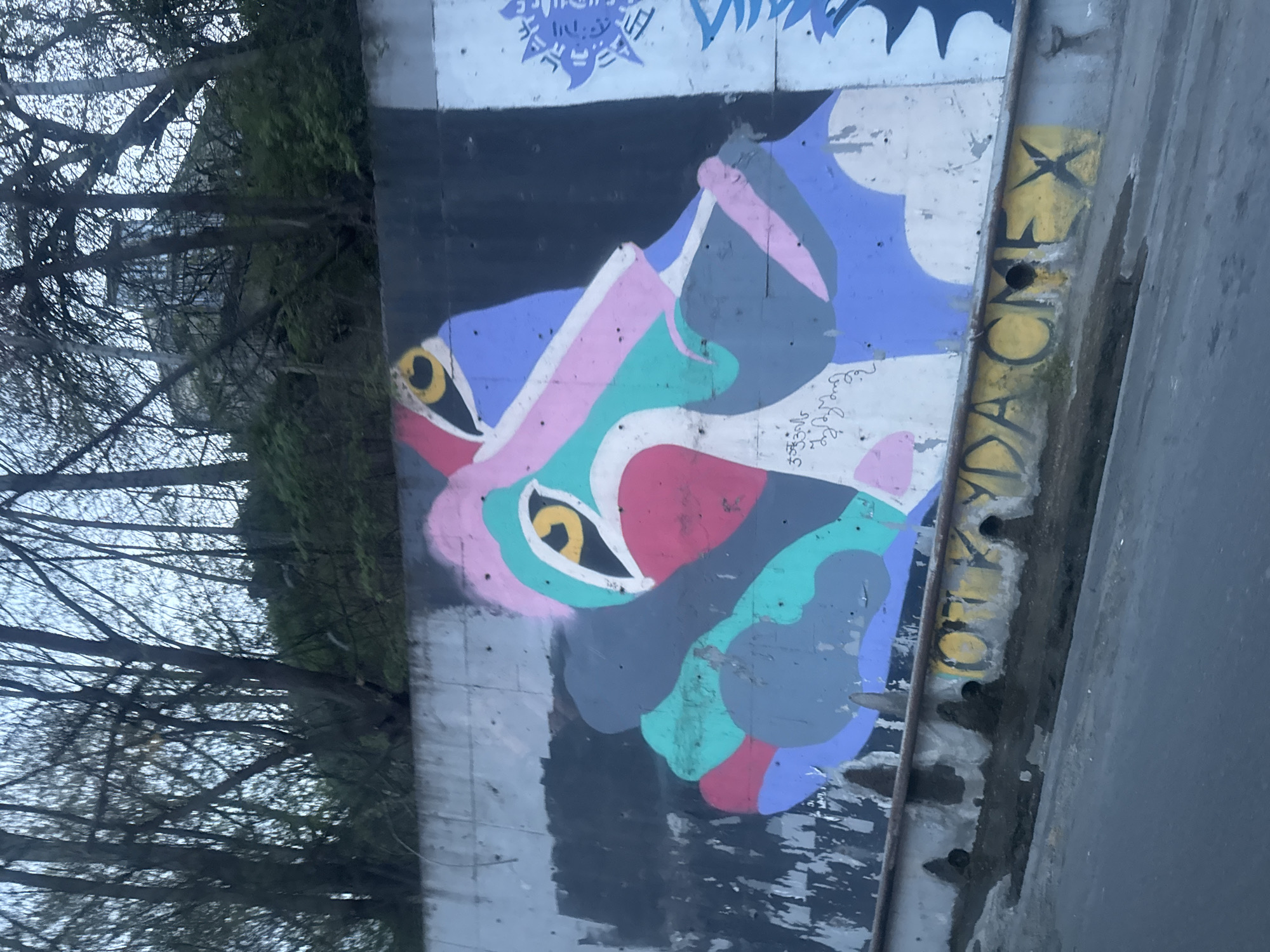 A concrete wall displaying street art is situated adjacent to a tree and a roadway. The art depicts a stylized image of animal faces in overlapping colorful shapes with prominent yellow eyes. The artwork includes traces of tagging and other markings on the wall. The location is identifiable as Tbilisi, Georgia based on the style of street art commonly found in the city.