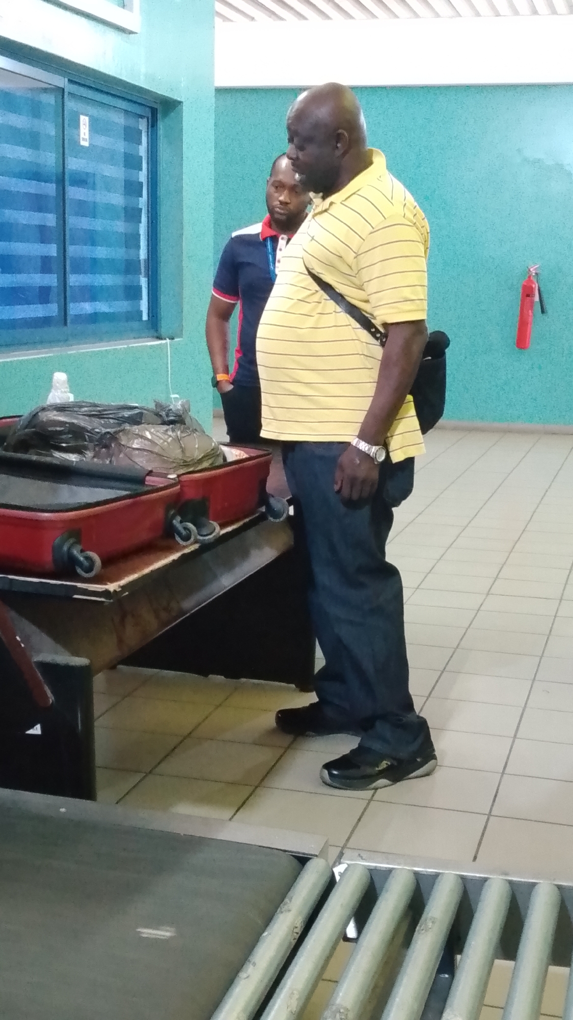 The image depicts two men standing near a luggage conveyor belt and baggage inspection table. The primary subject is an African male with a shaved head, wearing a yellow and white striped collared shirt, dark blue jeans, and black shoes with silver accents. He has a black shoulder bag and wears a watch. The second male, also African, is positioned behind him, wearing a navy blue polo shirt with red and white trim, and dark pants. A red fire extinguisher is visible on the wall behind them. A red suitcase with black bags on top sits on the baggage inspection table. The flooring is tiled in a light color. The location is identified as Orerokpe, Nigeria, implying the scene is likely at an airport or transportation hub within the city.