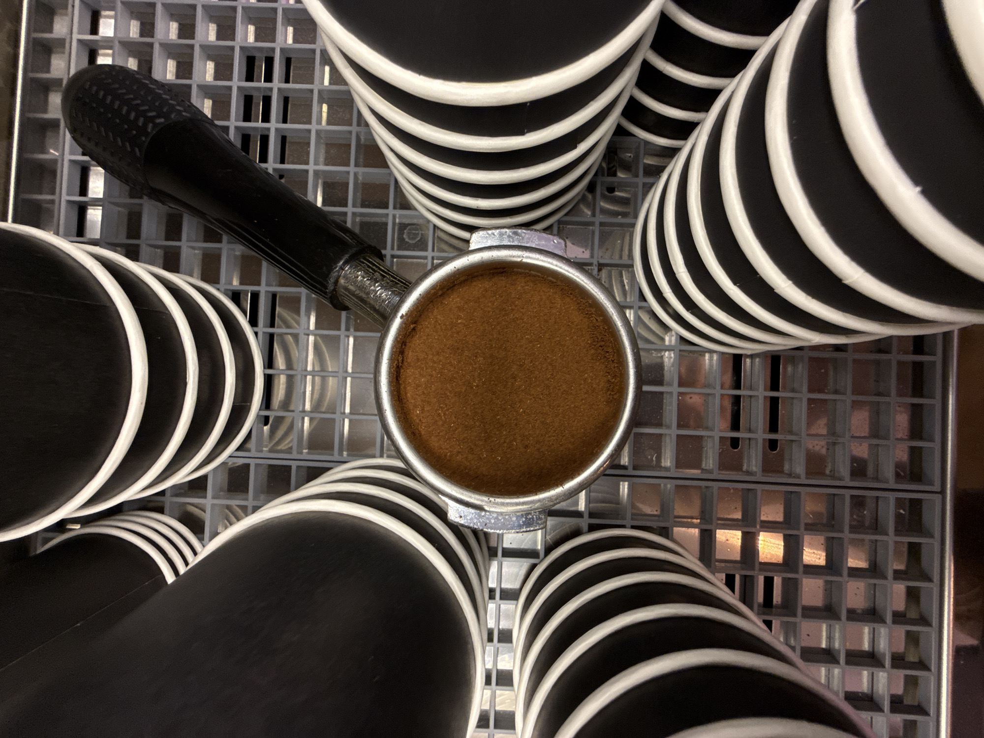 The image displays a close-up, top-down view of a portafilter filled with ground coffee, resting on a grey, grid-like surface. Surrounding the portafilter are stacks of black paper cups with white stripes. The scene appears to be indoors, likely in a cafe or kitchen environment, and is not specific to Dolgoprudny, Russia. There are no people, actions, or interactions depicted.