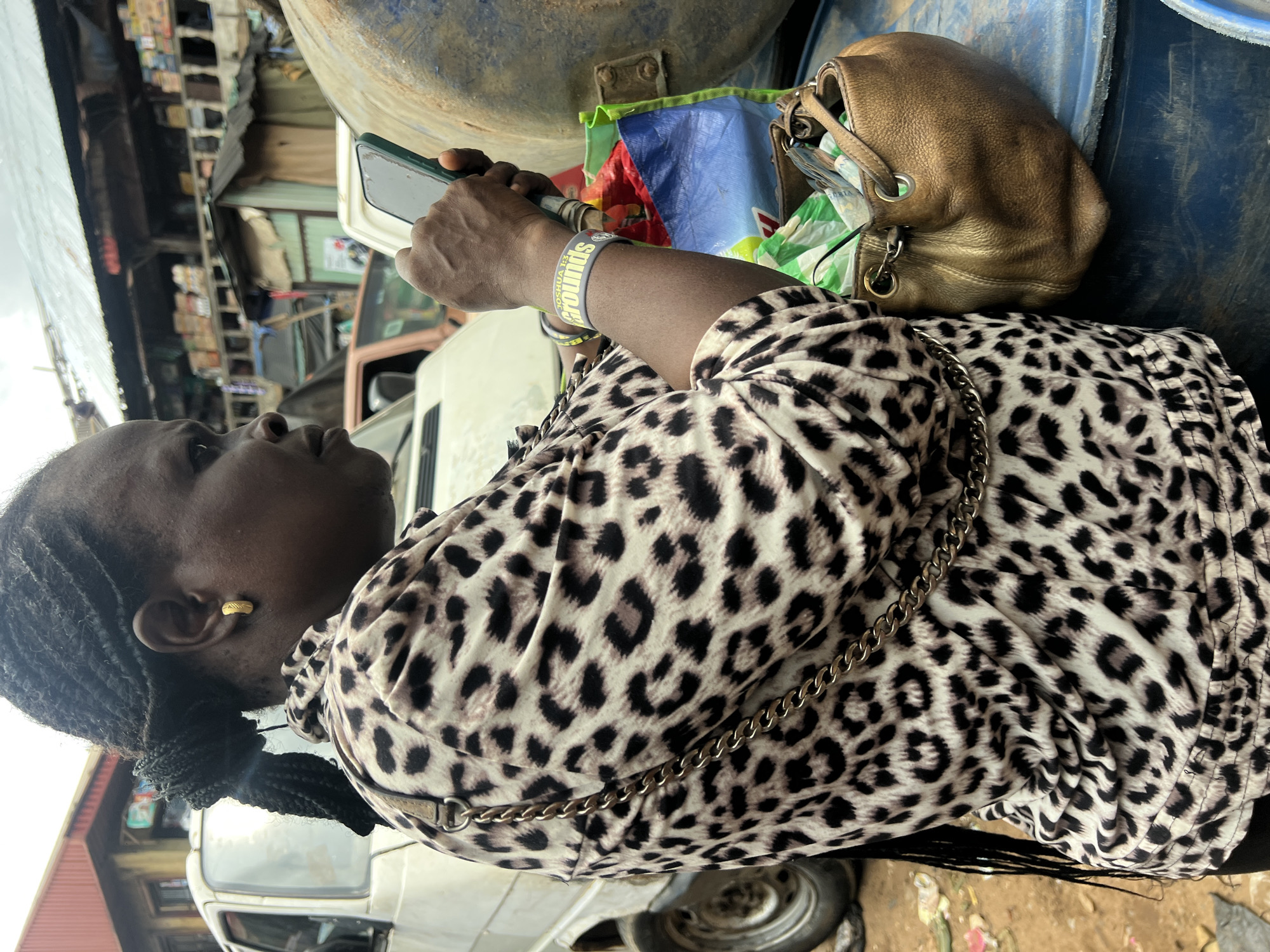 A medium shot captures a Black woman, viewed in profile from the right, engaged with her smartphone. She has her hair braided in a bun with a long braid extending down her back, and she wears a gold hoop earring in her left ear. Her clothing is a leopard-print blouse with a gold chain strap draped across it. On her left wrist, she wears a grey wristband with "JOSHUA 1:3 GROUNDS" visible. She holds a dark-colored smartphone in her right hand. Beside her, to the right, is a large, blue, plastic barrel, and resting on it is a small, metallic gold handbag containing various items, including what appears to be a blue and green reusable shopping bag. In the background, various structures, including a white van and what appear to be market stalls with corrugated metal roofs and shelves stocked with goods, suggest a bustling urban environment. The ground is littered with debris. The overall impression is of a moment captured in a busy marketplace.