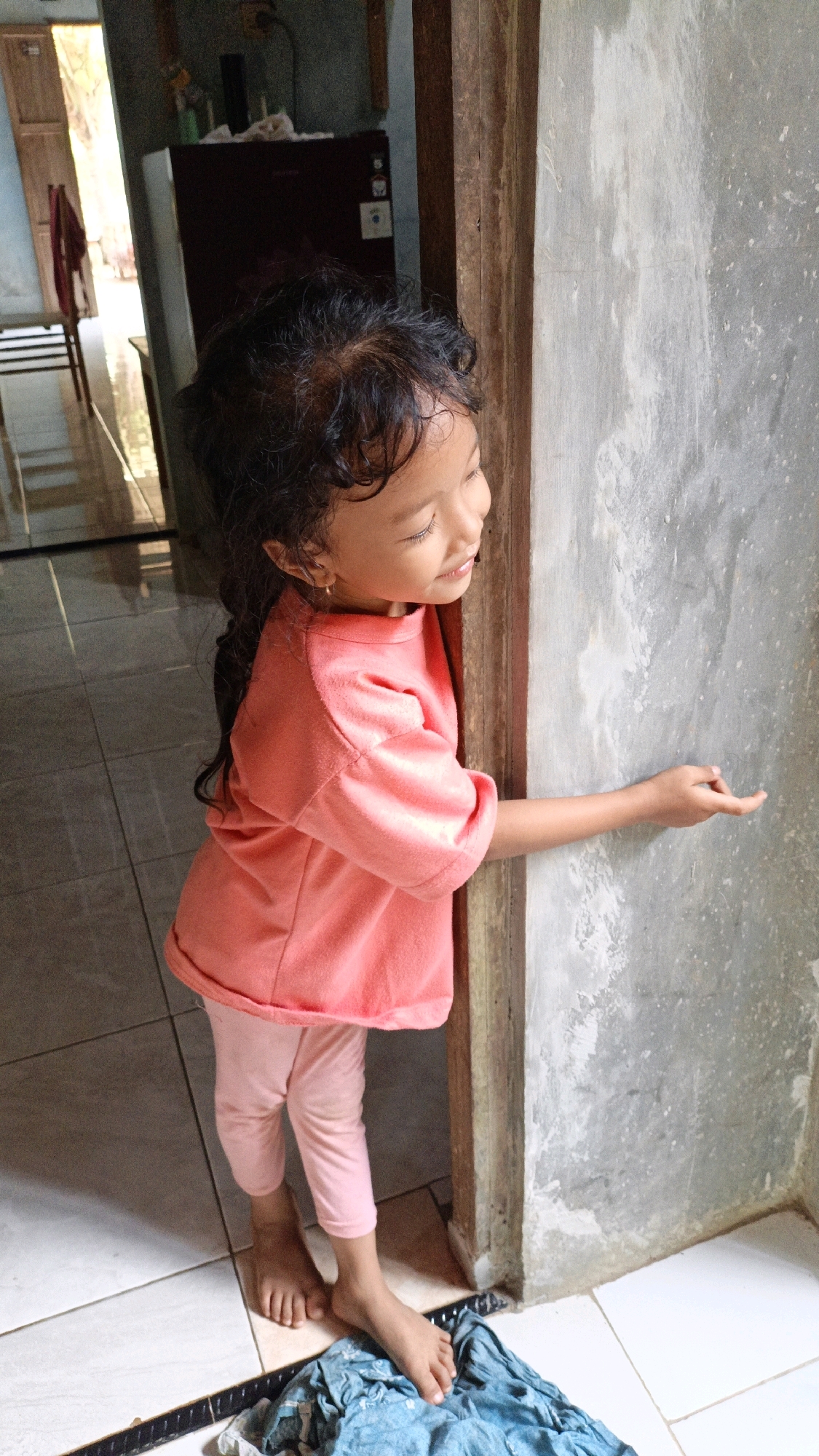 In Suwawal Timur, Indonesia, a young girl with dark, curly hair stands to the right of a doorway, facing outwards and looking to her right. She wears a pink short-sleeved shirt and pink leggings, with her bare feet visible on a tiled floor. Her right arm is extended, with her index finger pointing towards a rough, grey wall. The background shows an interior space with a maroon refrigerator and an open doorway leading to an outdoor area with wooden chairs and a red cloth hanging. The scene appears to be in a simple dwelling with tiled floors and concrete walls.