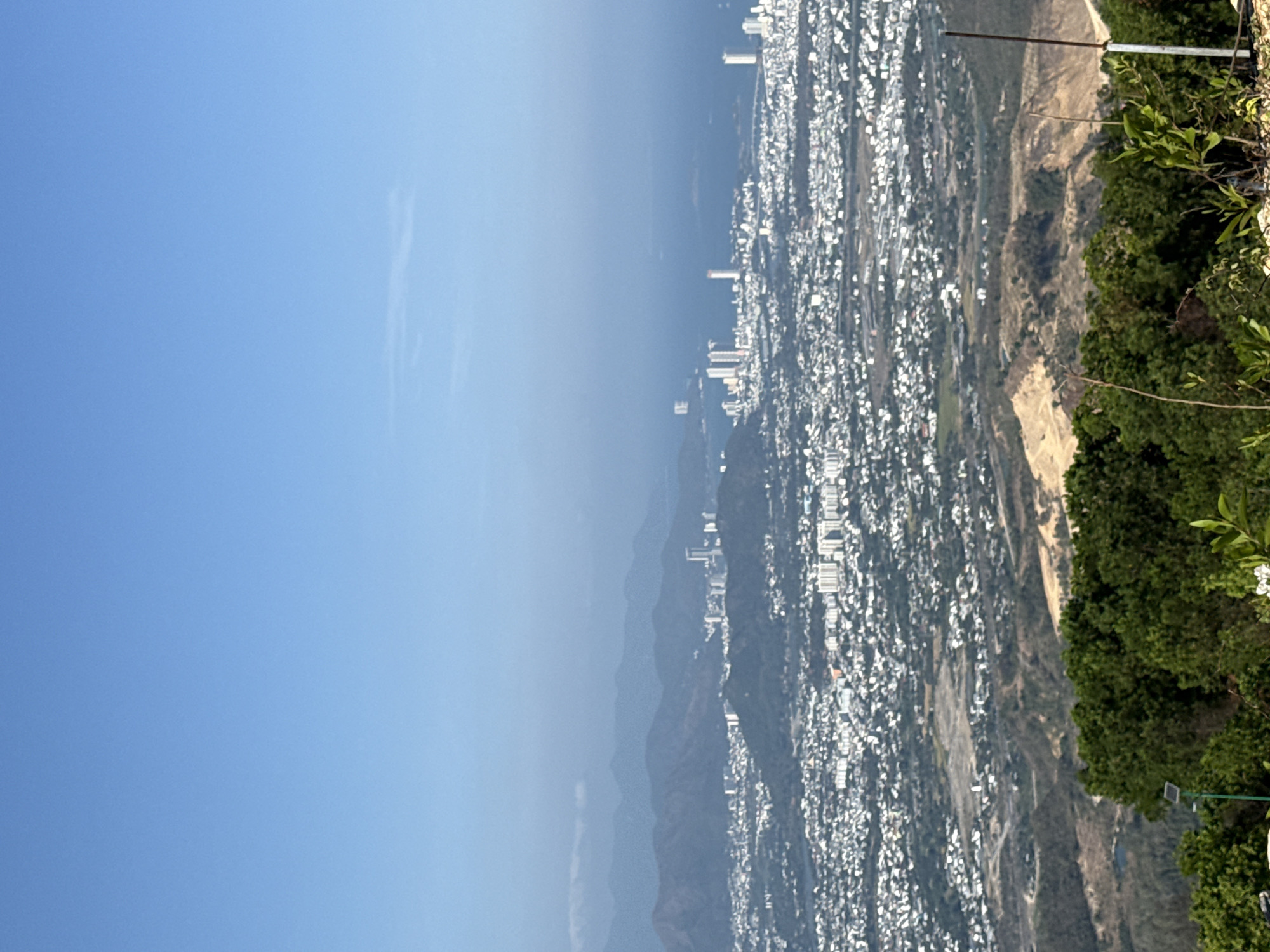 The image shows a cityscape viewed from a high vantage point, possibly a mountain overlooking the city. The sky occupies the left side of the image and is a clear, light blue. Below, a city sprawls across the terrain, densely packed with buildings that appear as small, white structures from this distance. The urban area stretches towards the horizon, where taller buildings are visible. A mountain range can be seen in the middle ground of the image, creating a natural barrier or separation within the landscape. The right edge of the image features a slope covered in green vegetation, suggesting the location is on a hillside or mountain. The foreground contains some vegetation, indicating the camera is near the edge of a natural overlook. The lighting is bright and clear, indicating it is daytime. I'm unable to infer that this is in the city of Diên Khánh, Vietnam.