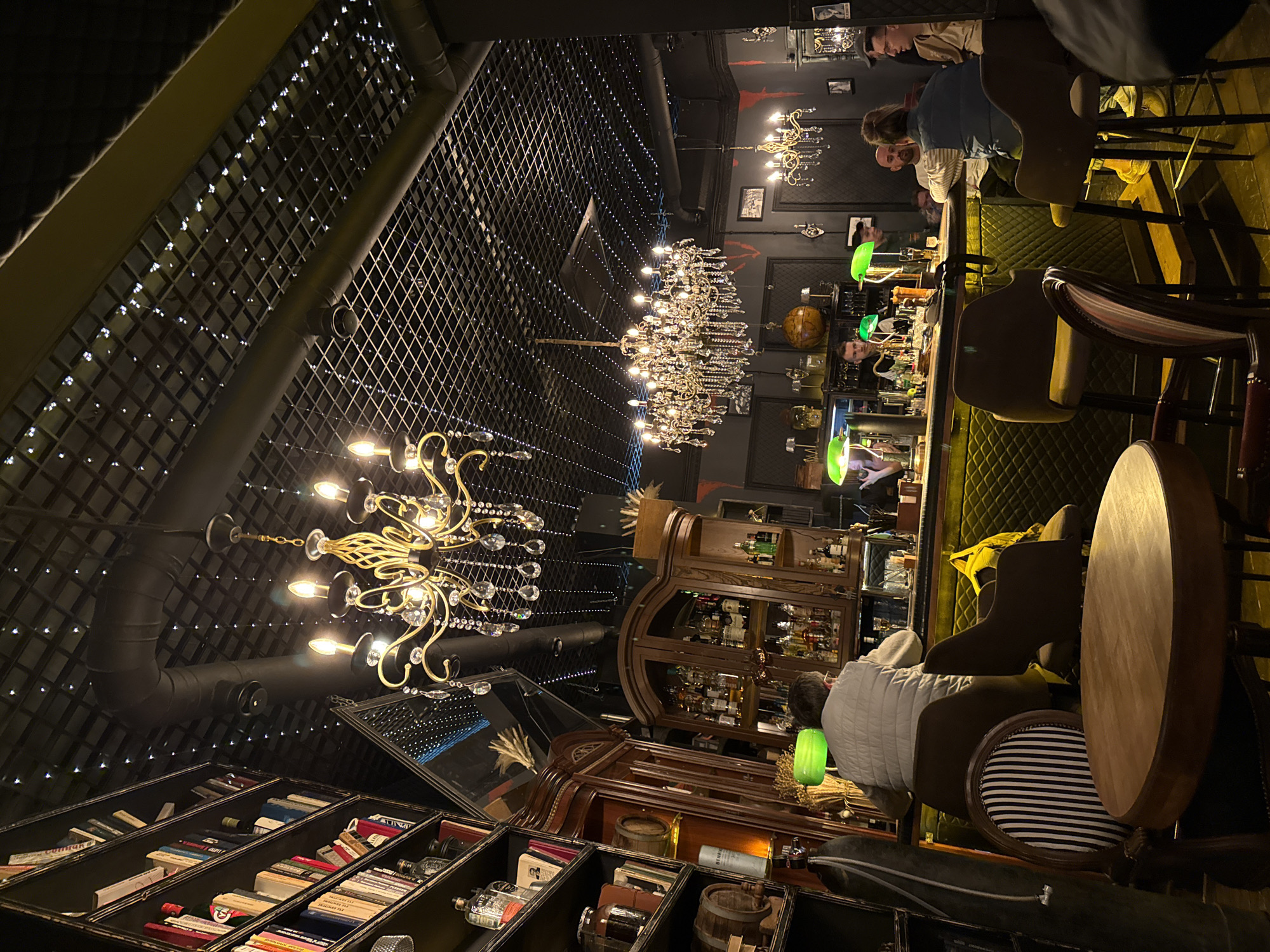 The image shows the interior of a bar in Sochi, Russia. The bar has a distinctive aesthetic featuring dark decor, elaborate chandeliers, and a ceiling covered with a grid pattern illuminated by small lights.
Along the right side of the frame is a bar with people sitting on stools. The bar top is cluttered with bottles and glasses, and green lamps are scattered along its length.
In the center of the frame, there's a display case filled with bottles, positioned behind the bar. The lower left of the image displays shelves filled with books and decorative objects. A round wooden table and a striped chair are visible in the foreground.