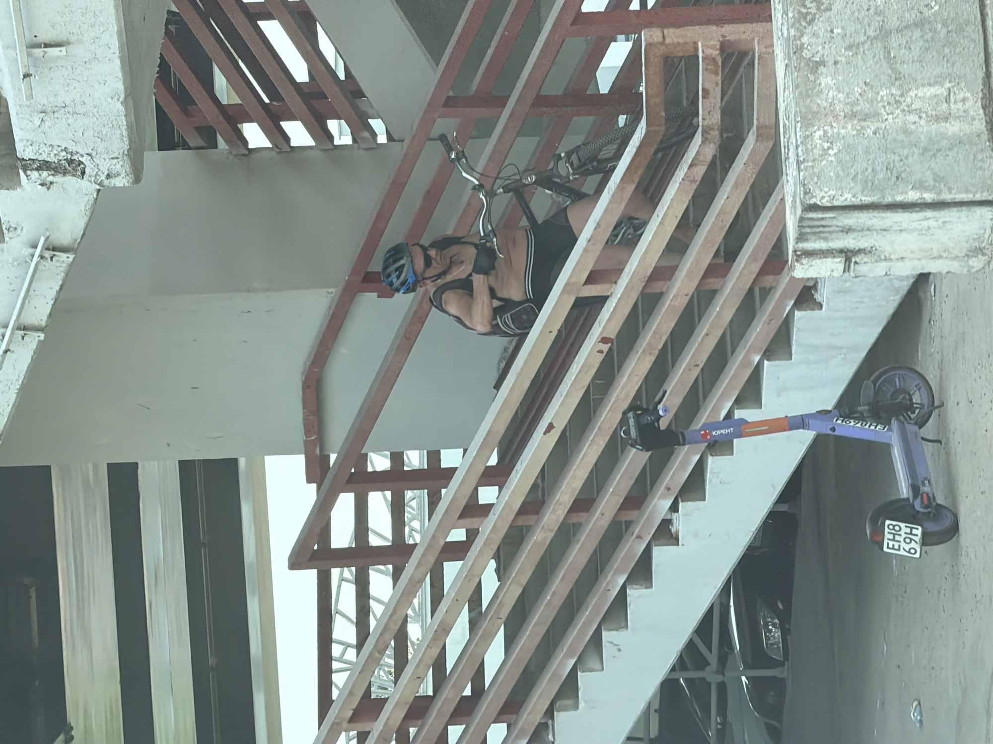 A man with a helmet is carrying a bicycle down a metal staircase. The stairs appear to be part of an overpass or elevated walkway. To the right of the stairs, an electric scooter is parked. The scooter has a license plate that reads "EH8 69H." The text "ЮРЕНТ" is visible on the scooter. This suggests that the scene is likely in Moscow, Russia, where "ЮРЕНТ" is a known electric scooter sharing service.