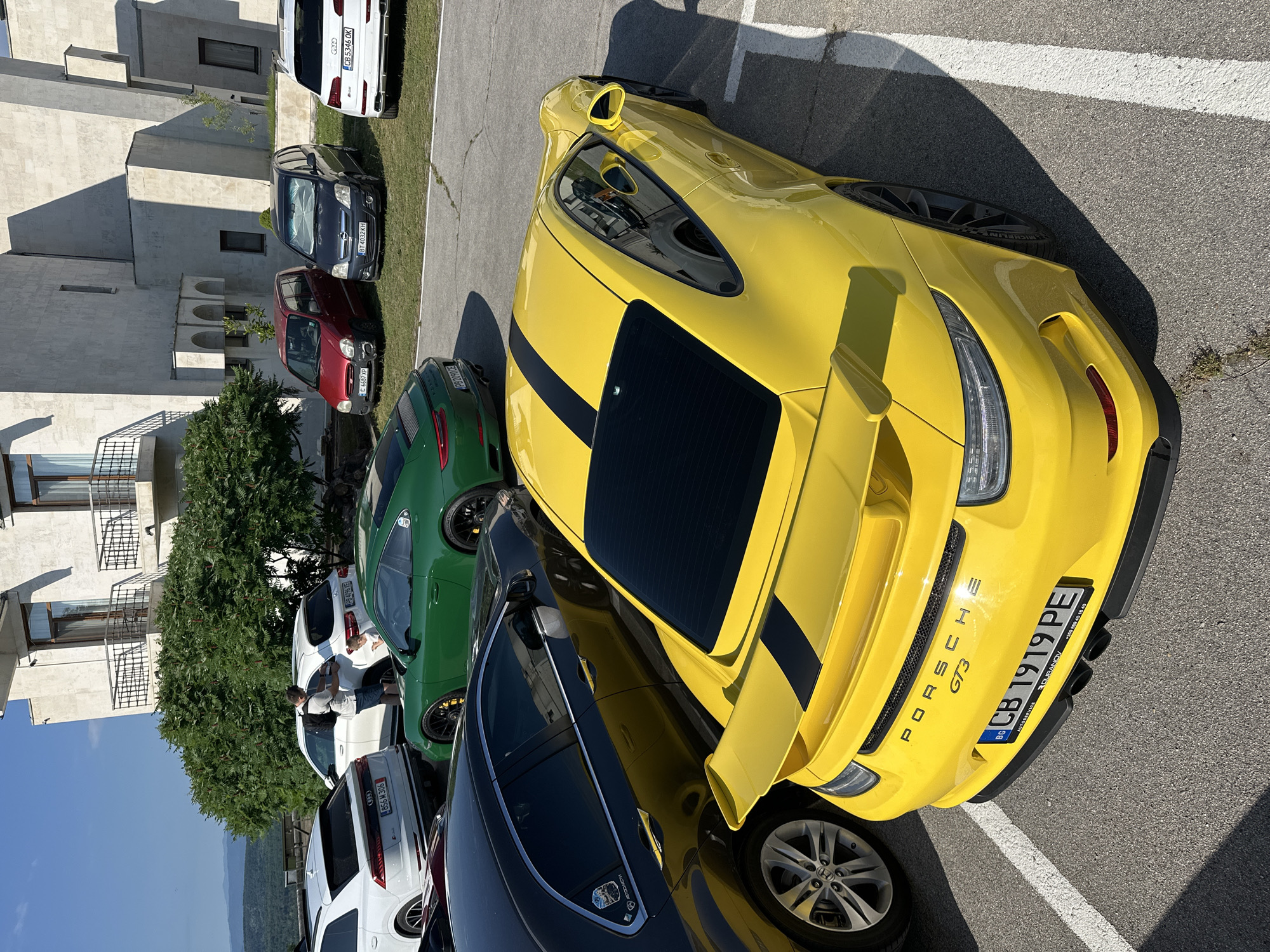 A bright yellow Porsche GT3 is parked in a lot with cracked asphalt, marked by white lines. The car has black racing stripes on the hood and roof, and a large rear spoiler. Its license plate reads "BG CB 1919 PE". In the background, several other cars are visible, including a green Porsche, a black sedan, a white Audi SUV, and a red hatchback. A white Mercedes-Benz SUV is also parked nearby. The scene appears to be in Arbanassi, Bulgaria, with a large building with balconies and arched windows in the background, and a lush green landscape beyond. The sky is clear and blue.
