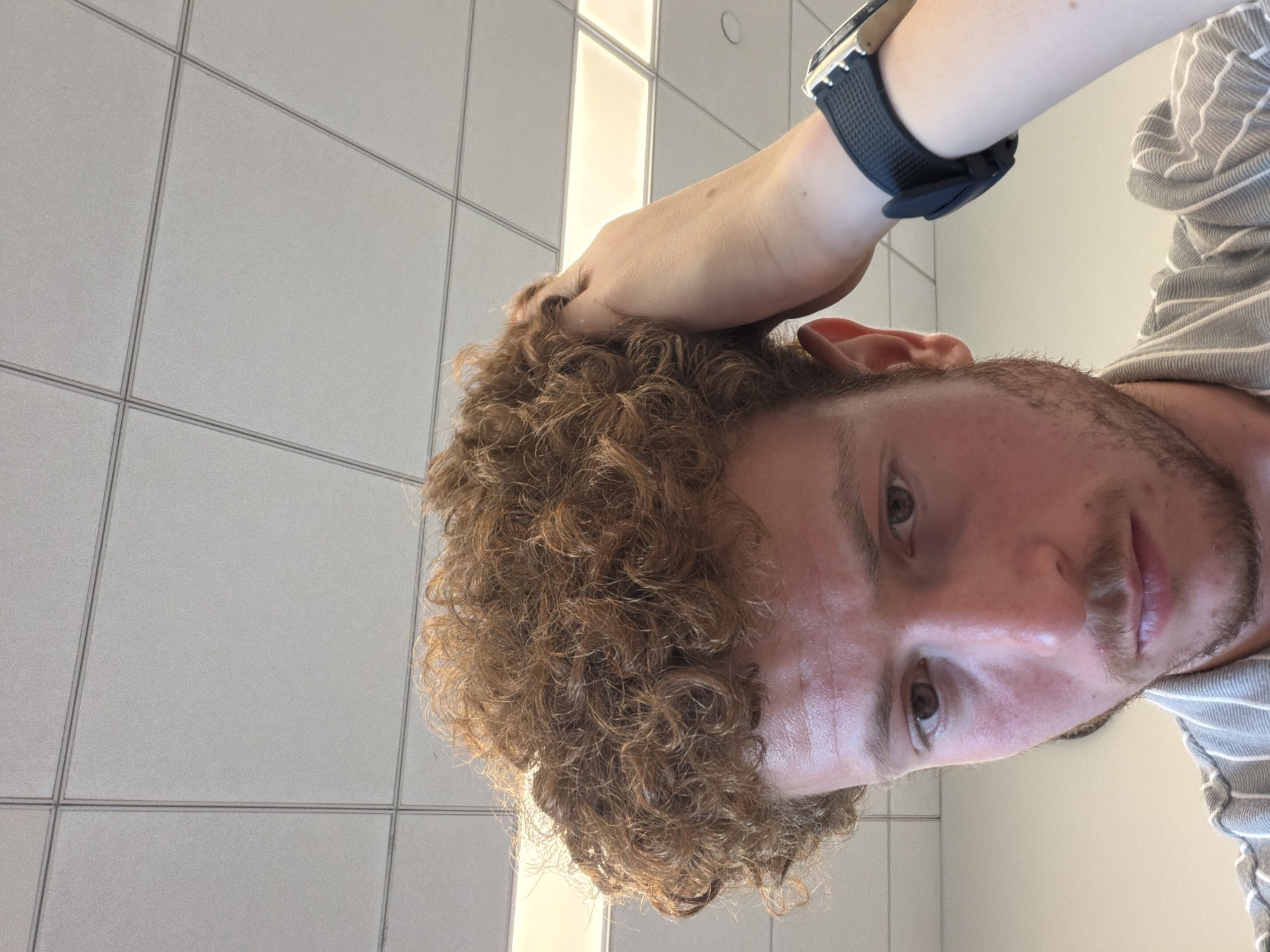 A close-up, rotated portrait shot depicts a man with curly, reddish-brown hair, fair skin, and a trimmed beard. He is wearing a black wristwatch on his left wrist, and a gray and white striped shirt is partially visible. His left hand is touching his hair. The background consists of a grid of white tiles. The location is not discernible from the image.