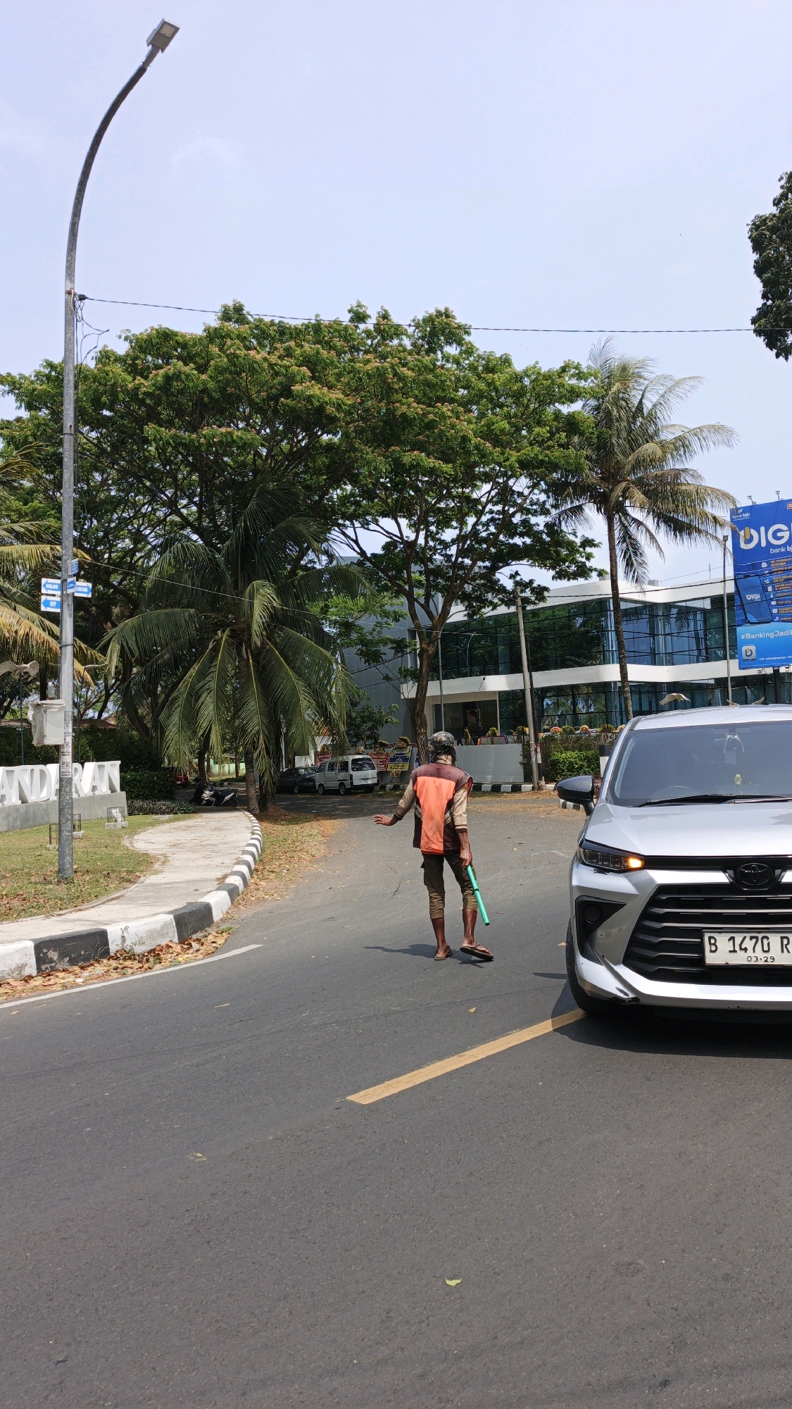 In Desa Wonoharjo, Indonesia, a person wearing an orange vest and brown pants is standing on an asphalt road, holding a green broom. The person is barefoot and appears to be directing traffic or performing sweeping duties. A silver car with a license plate reading "B 1470 R" is parked on the right side of the road, its front bumper is visible. In the background, there is a modern building with large glass windows and several palm trees and other lush green trees. A street light stands on the left side of the image. The sky is clear and blue. The road has a yellow line painted in the center.